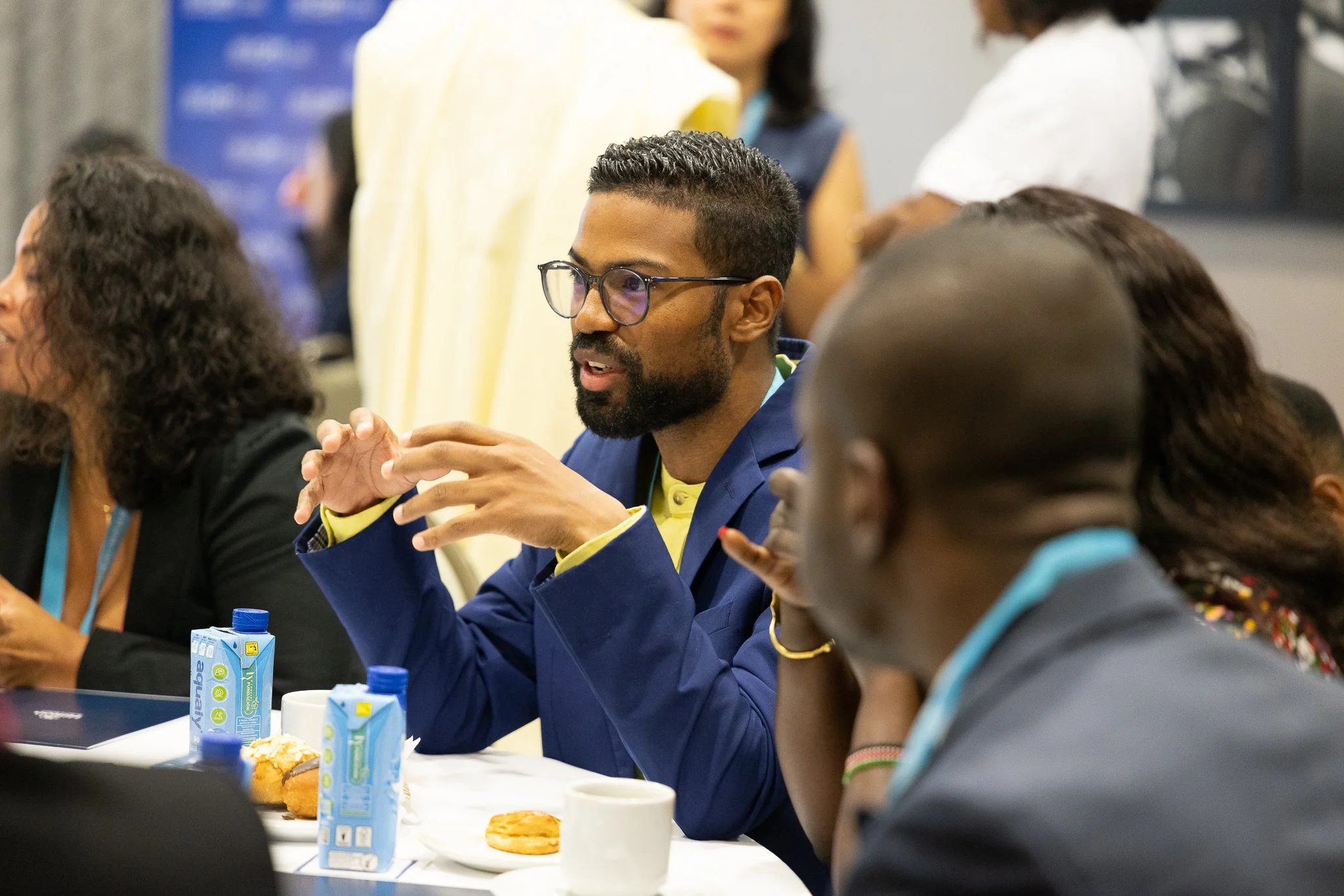 A man with glasses and a beard, wearing a blue blazer, is sitting at a table during a meeting or conference. He appears to be speaking or explaining something with hand gestures. Other people are seated around him, with some items like juice boxes, c