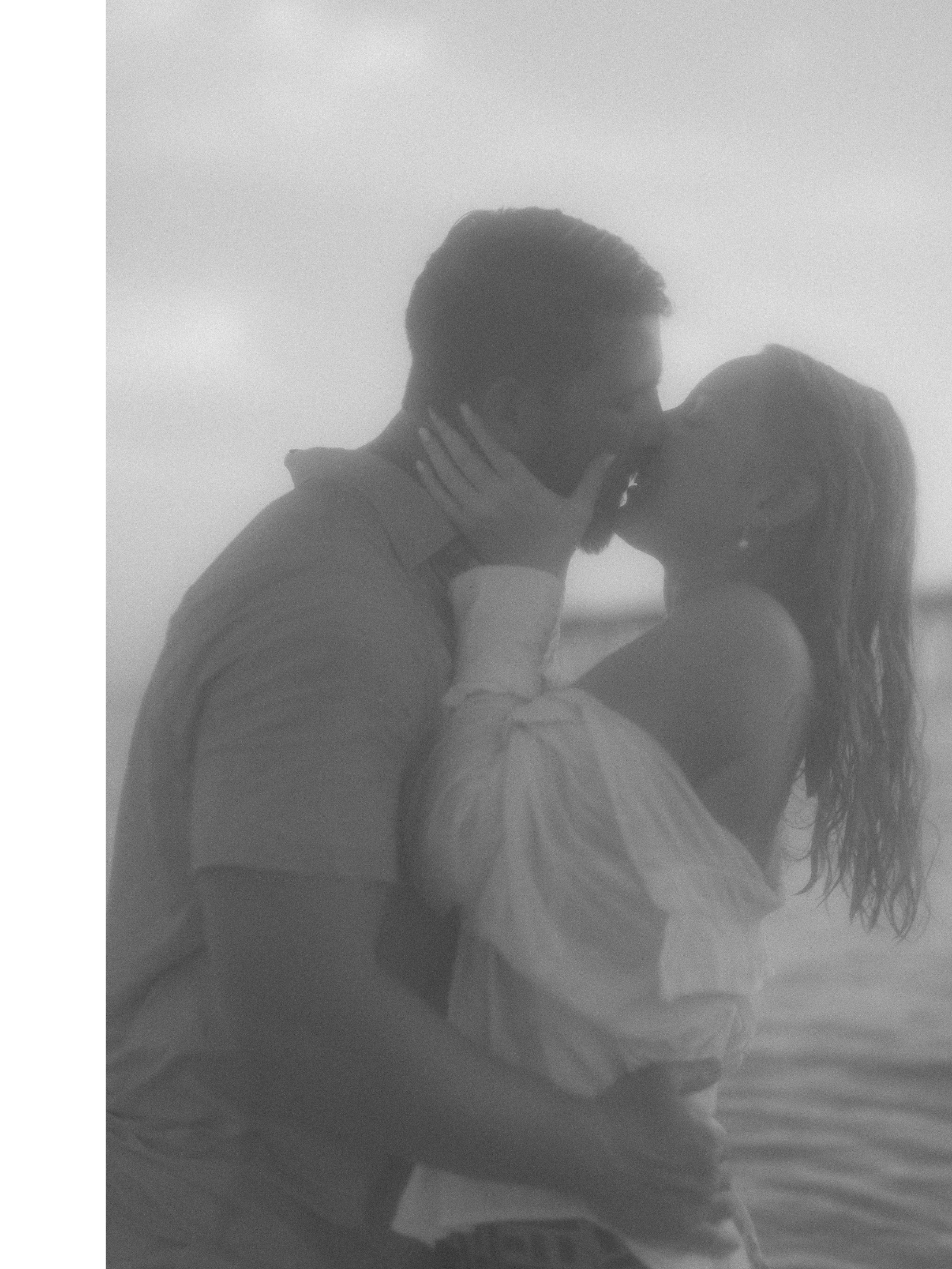 A black and white photo of a couple kissing on the beach, with the man holding the woman and her arms wrapped around his neck.
