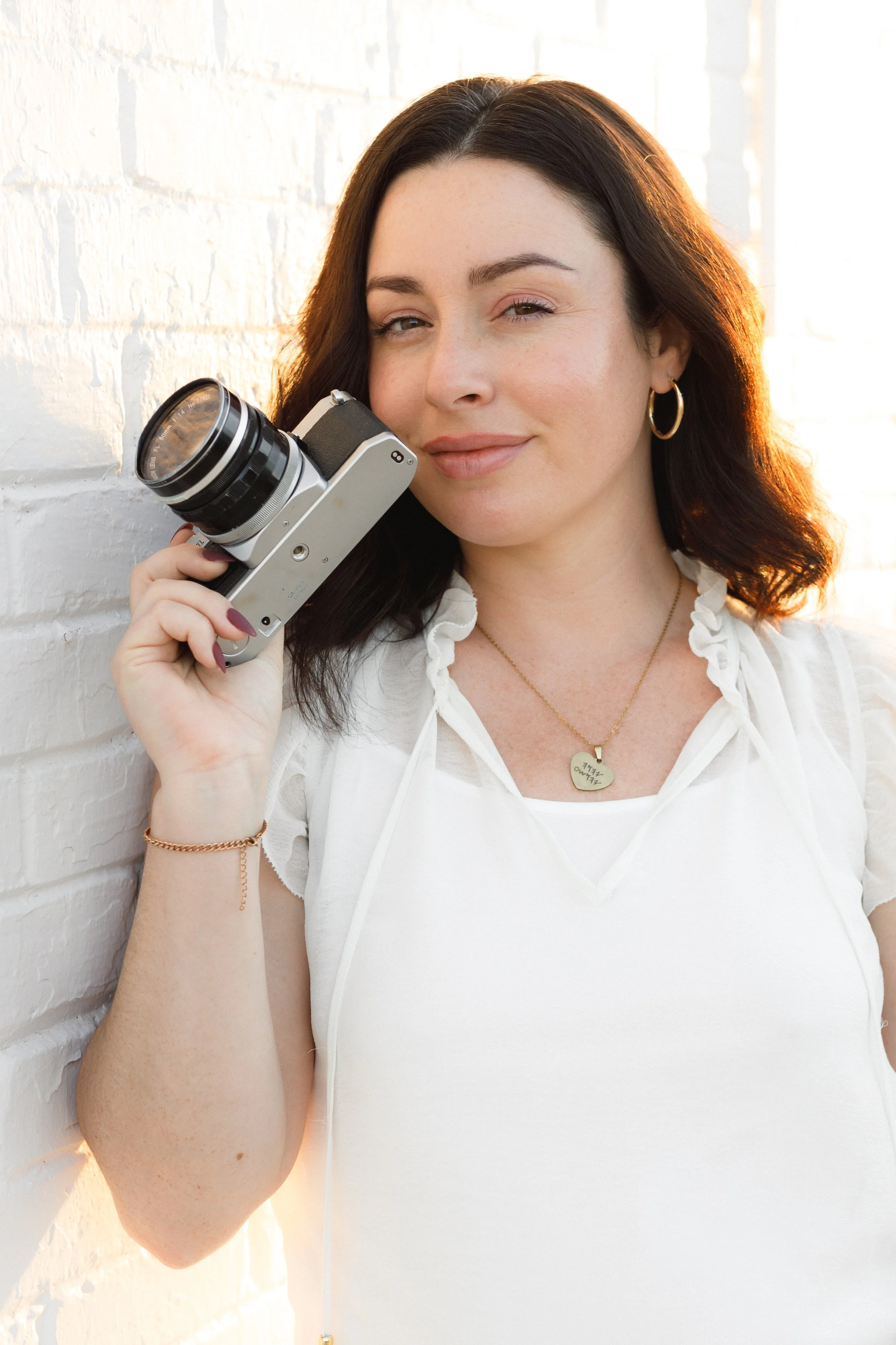 A woman with dark brown hair holding a vintage film camera to her face, standing against a white brick wall with warm sunlight.