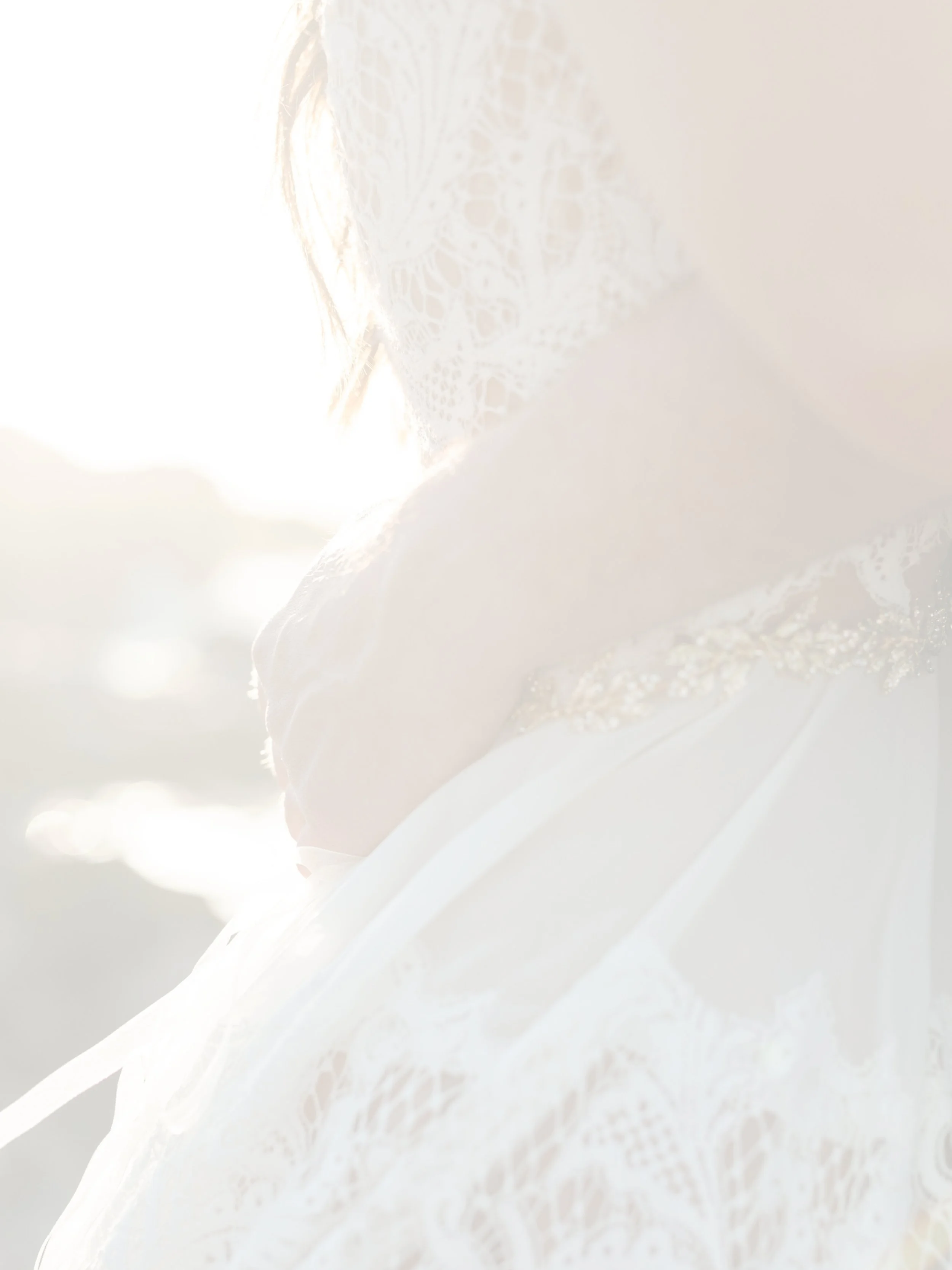 A woman wearing a white lace dress, partially illuminated by bright sunlight, with focus on her shoulder and part of her face. The background is overexposed with soft, glowing light.