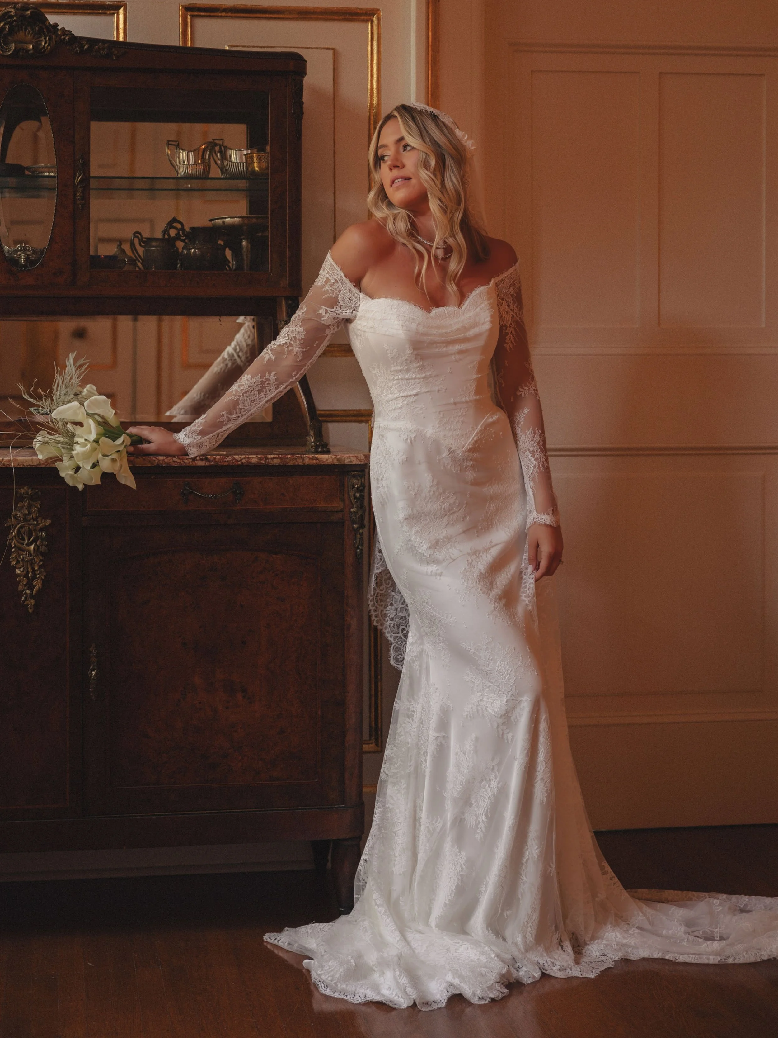 A woman in a white lace wedding dress standing in a vintage room with wooden furniture and gold framed wall decor, holding a bouquet of white flowers.