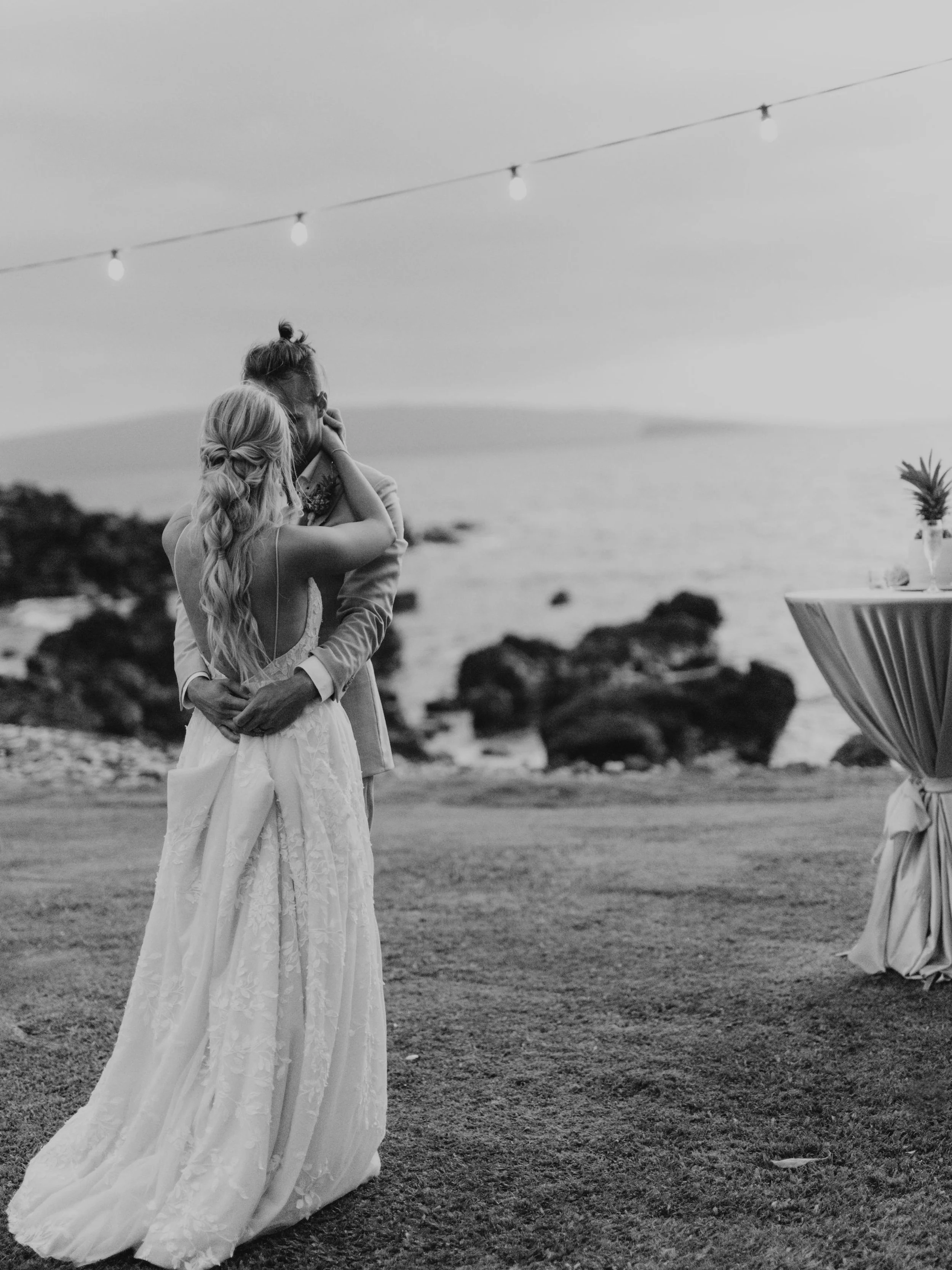 A bride and groom embrace at a seaside wedding, with rocks and water in the background, and string lights overhead.