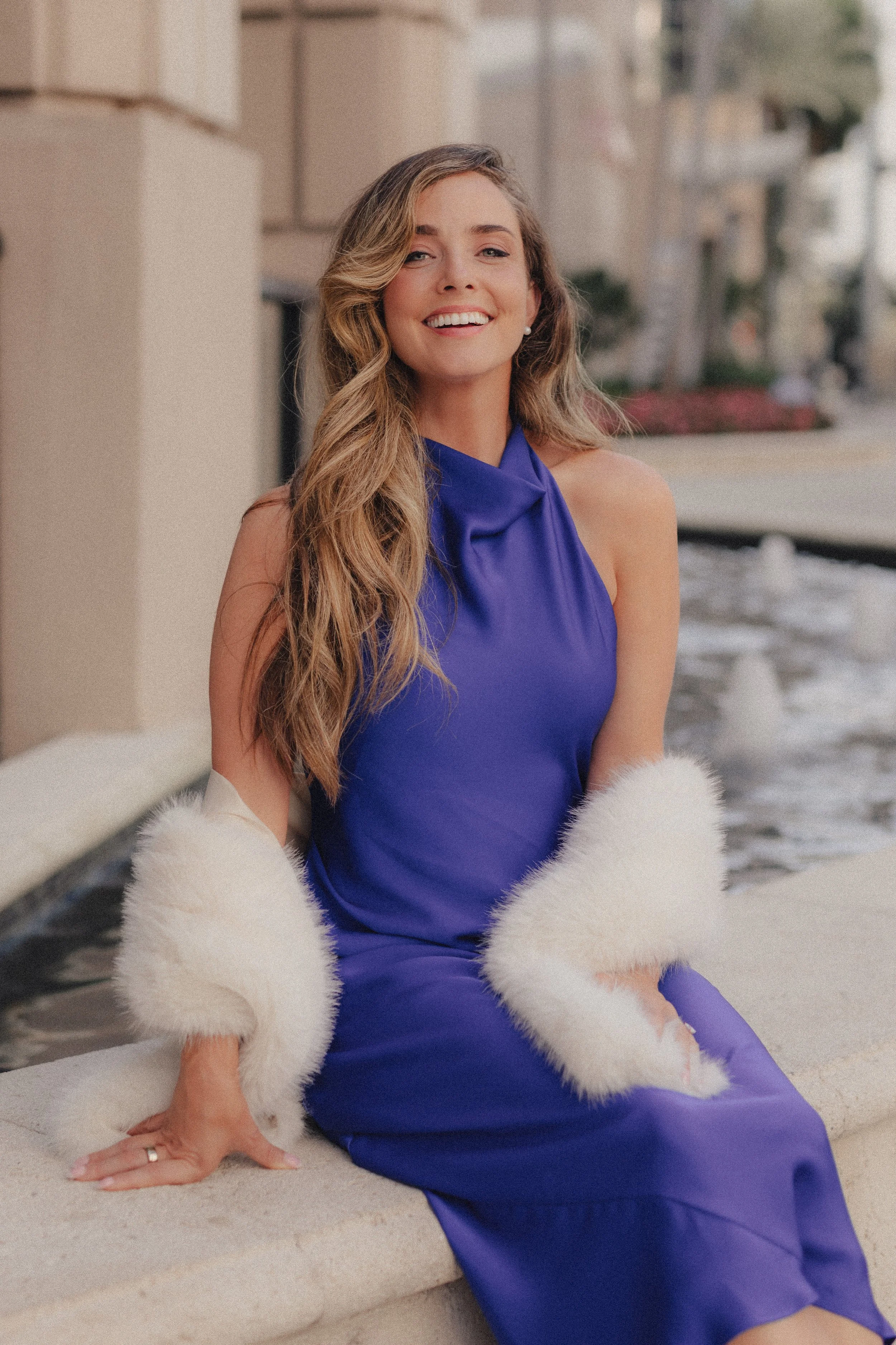 A woman in a blue satin dress and white fur stole sitting outdoors near a water feature, smiling at the camera.