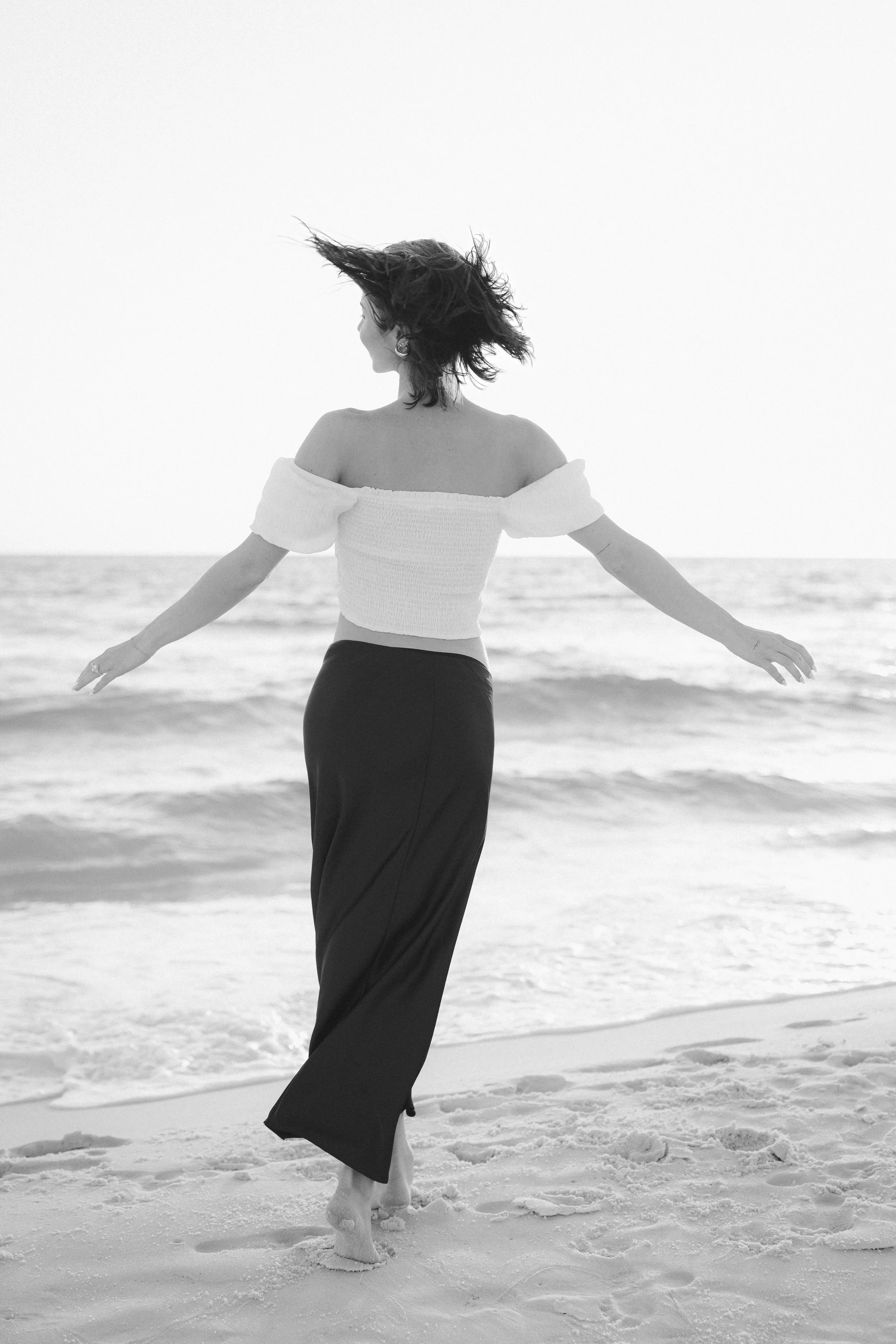 A woman with short hair by the ocean, standing barefoot on the sand, wearing a white off-the-shoulder top and long black skirt, with her arms extended outward.