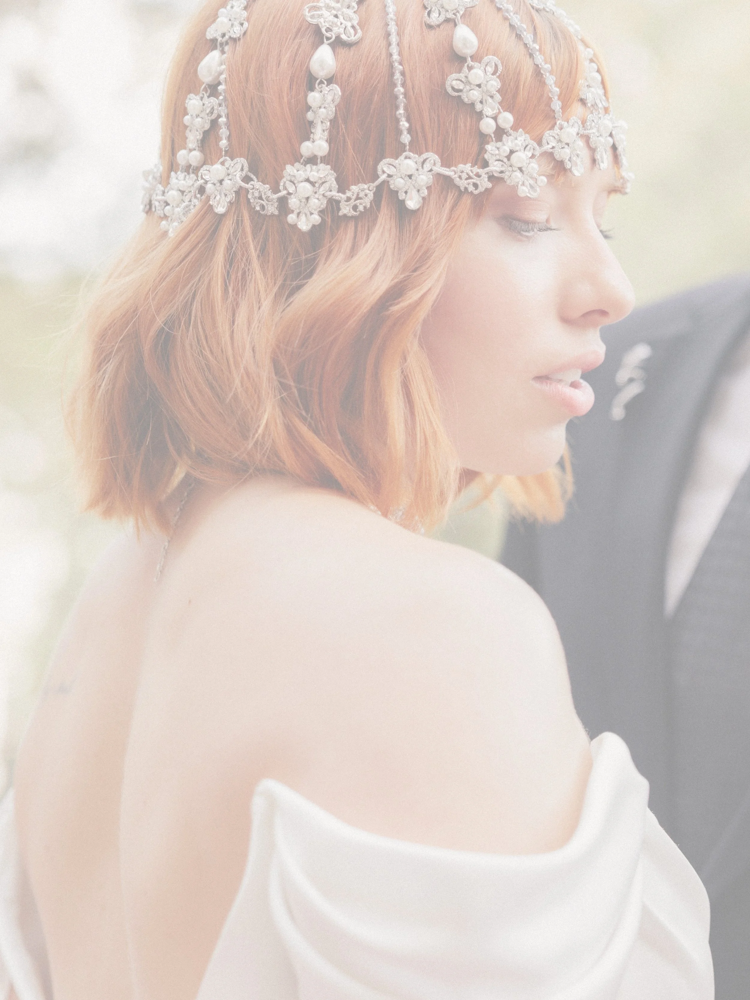 A woman with shoulder-length red hair, wearing a pearl and floral headpiece, looking to the side, dressed in a white off-the-shoulder garment, with a partly visible man in a dark suit behind her.