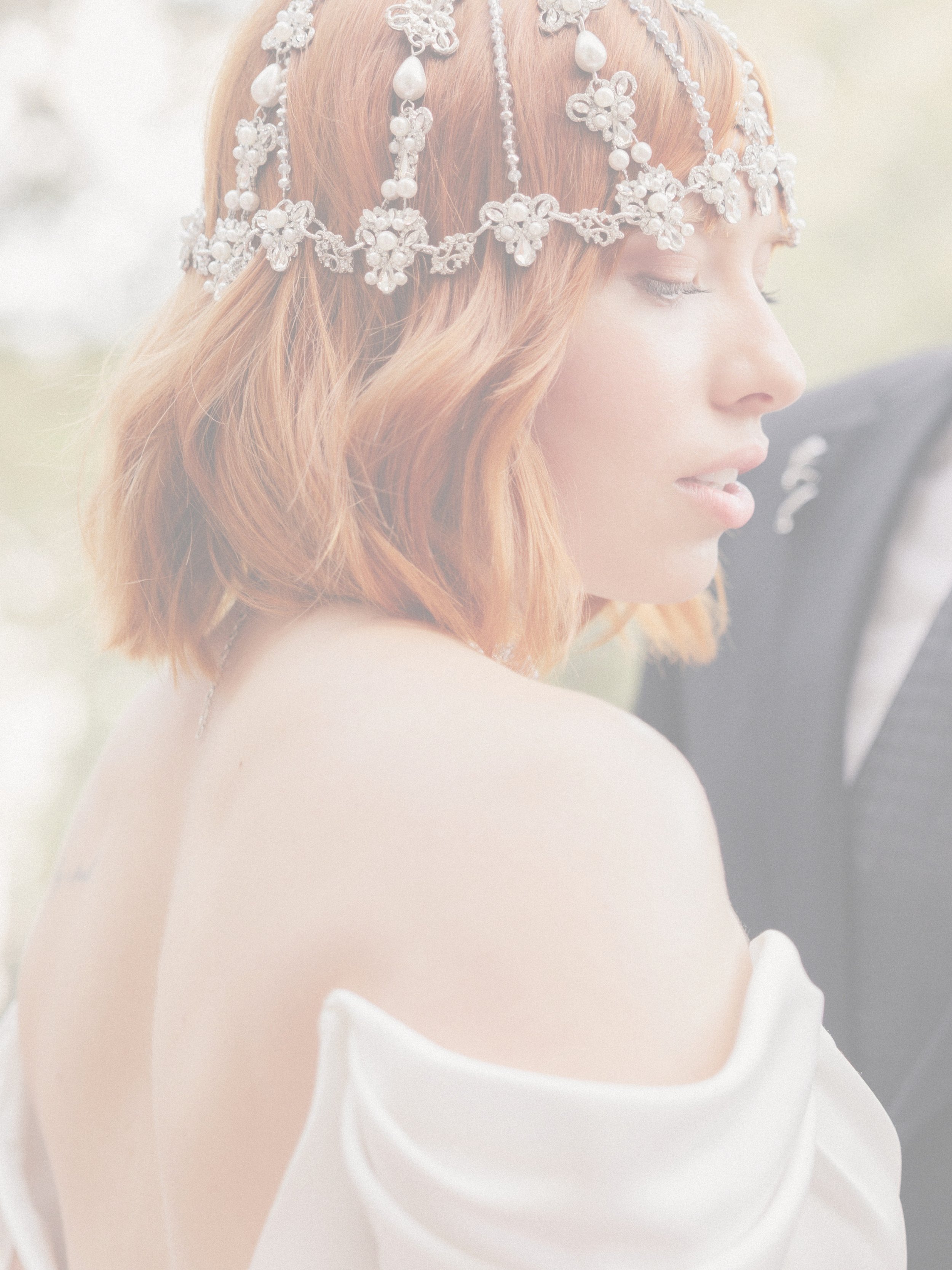 A woman with shoulder-length red hair, wearing a pearl and floral headpiece, looking to the side, dressed in a white off-the-shoulder garment, with a partly visible man in a dark suit behind her.