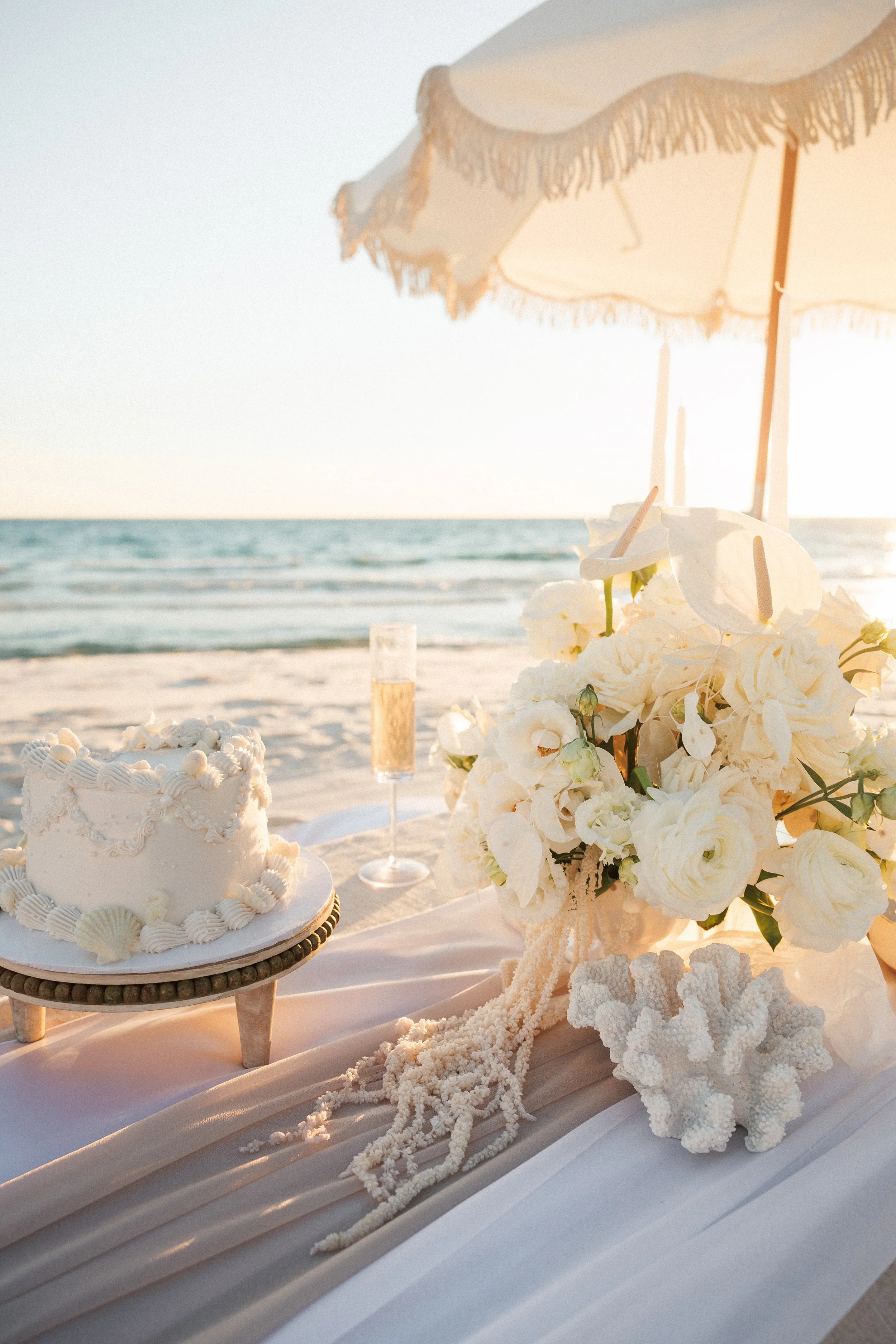 Wedding beach setup with a white cake on a stand, a bouquet of white flowers, a coral piece, and a champagne glass under a beige umbrella on the sandy beach during sunset.