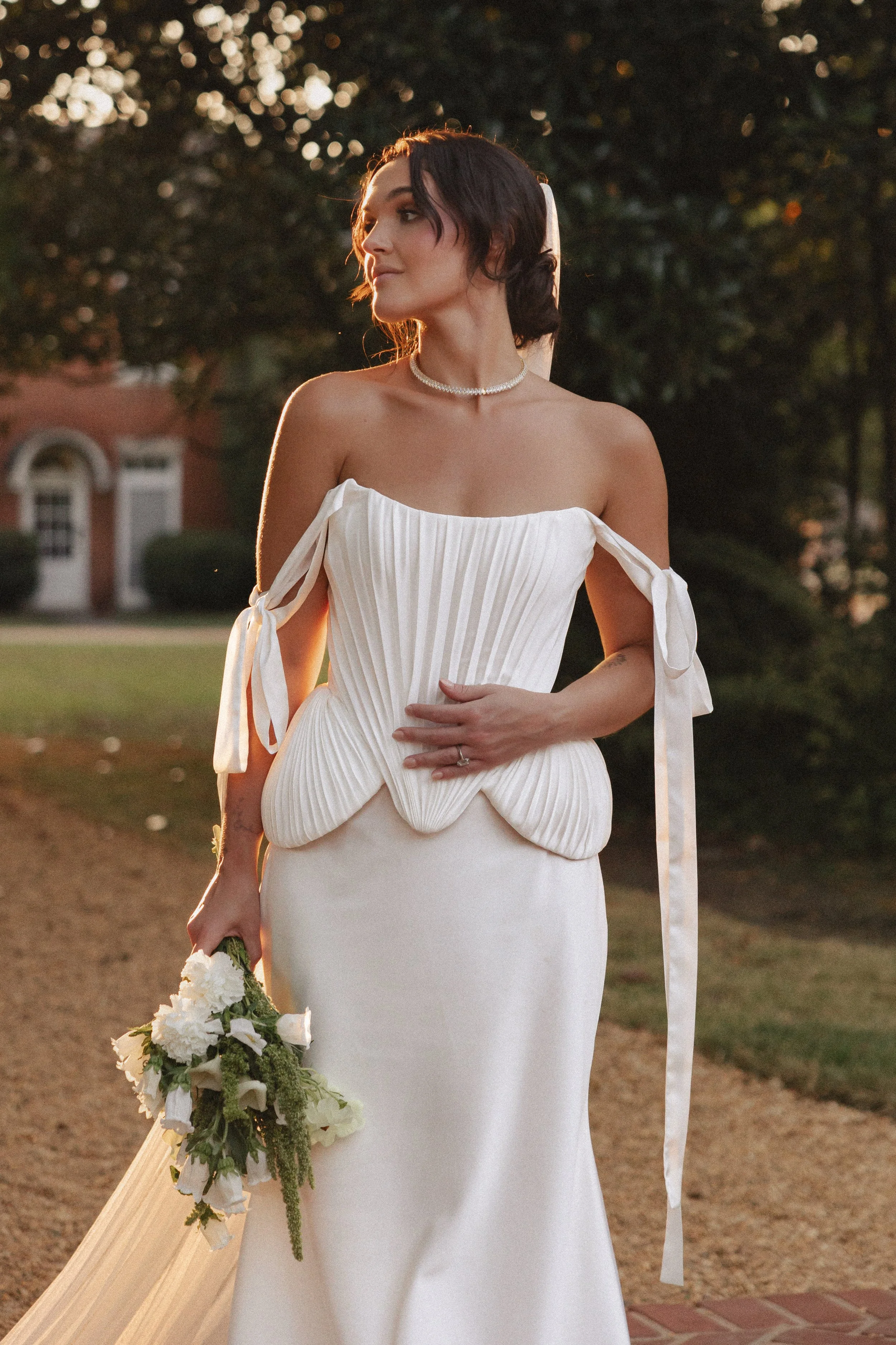 A woman in a white strapless wedding dress with tied ribbon sleeves, holding a bouquet of white flowers, outdoors during sunset.