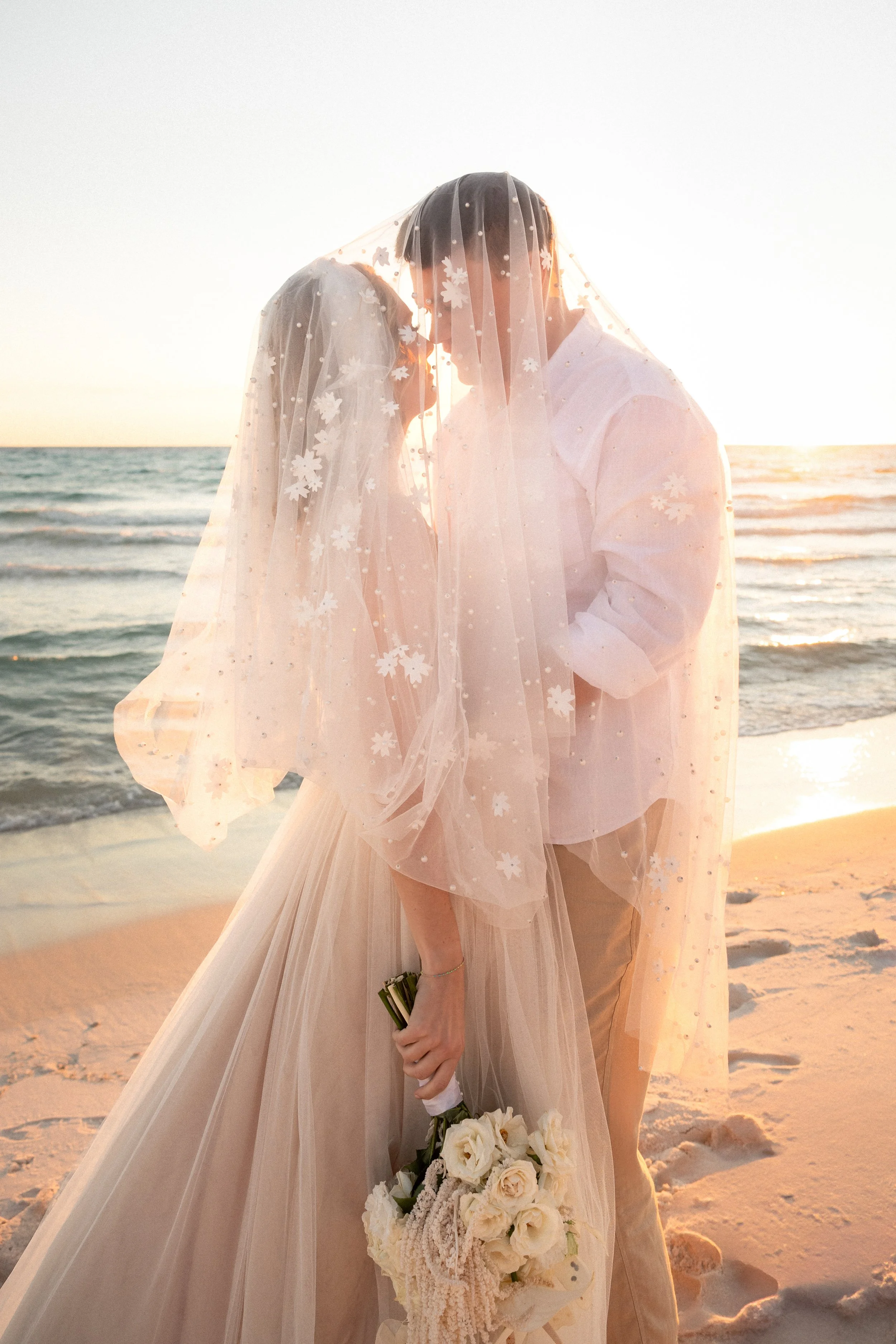 A couple dressed in wedding attire standing on a beach during sunset, with their foreheads touching and a bouquet of flowers in the woman's hand.