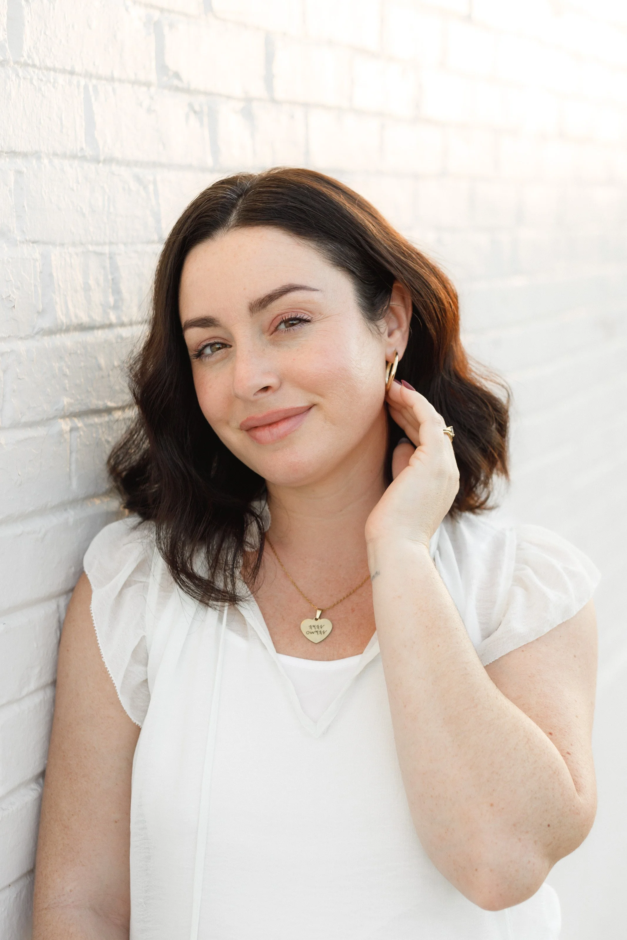 A woman with shoulder-length dark hair, wearing a white top and gold jewelry, poses against a white brick wall, smiling softly.