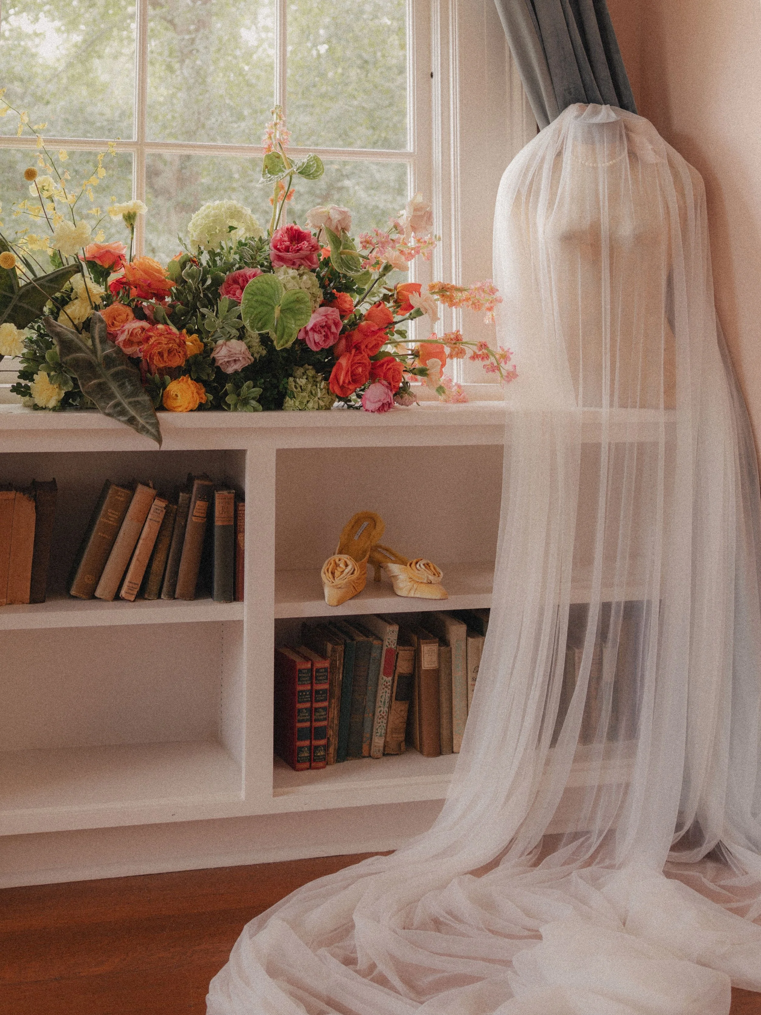 A white bookshelf with books and a pair of yellow high-heeled shoes, topped by a large bouquet of colorful flowers on a window sill, with sheer curtains and a window with a view of green trees outside.