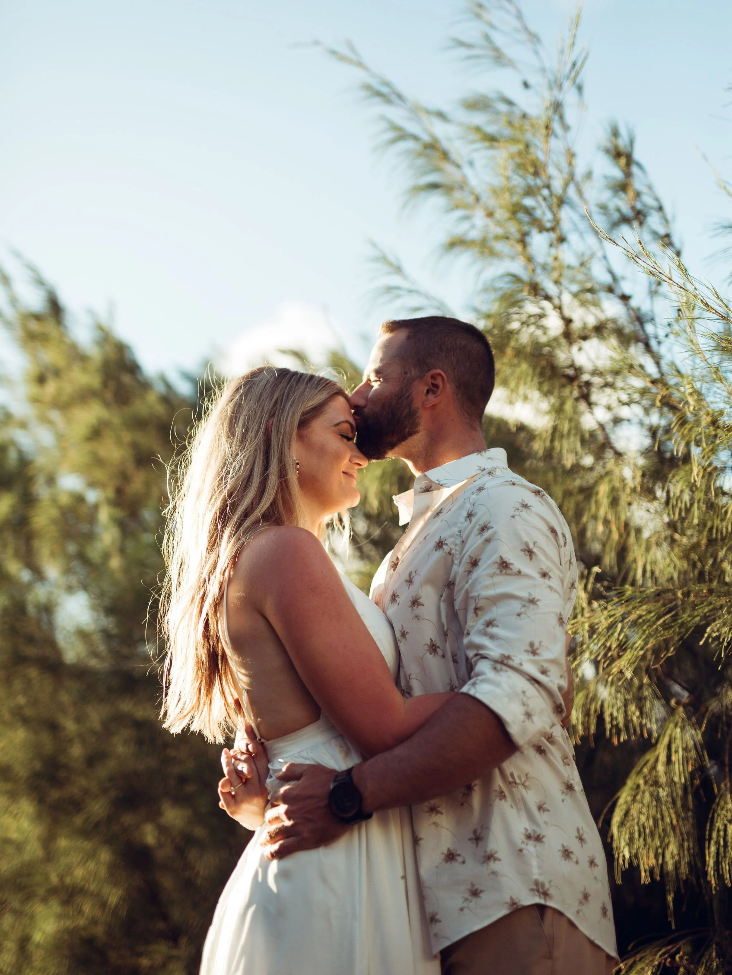 A couple in wedding attire sharing an intimate moment outdoors during sunset, surrounded by trees.