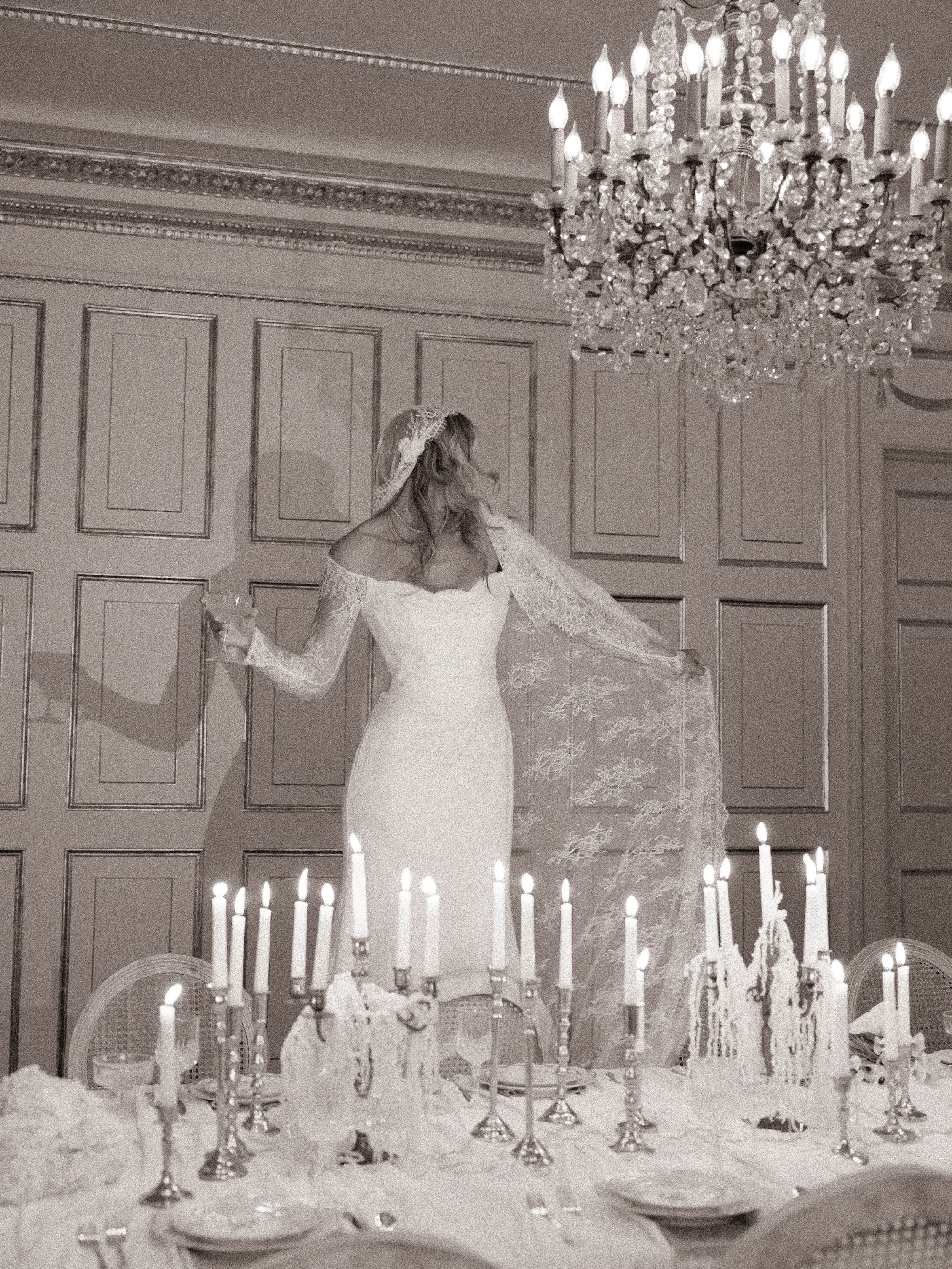 A woman in a wedding dress standing at a decorated dining table with candles and a chandelier overhead.
