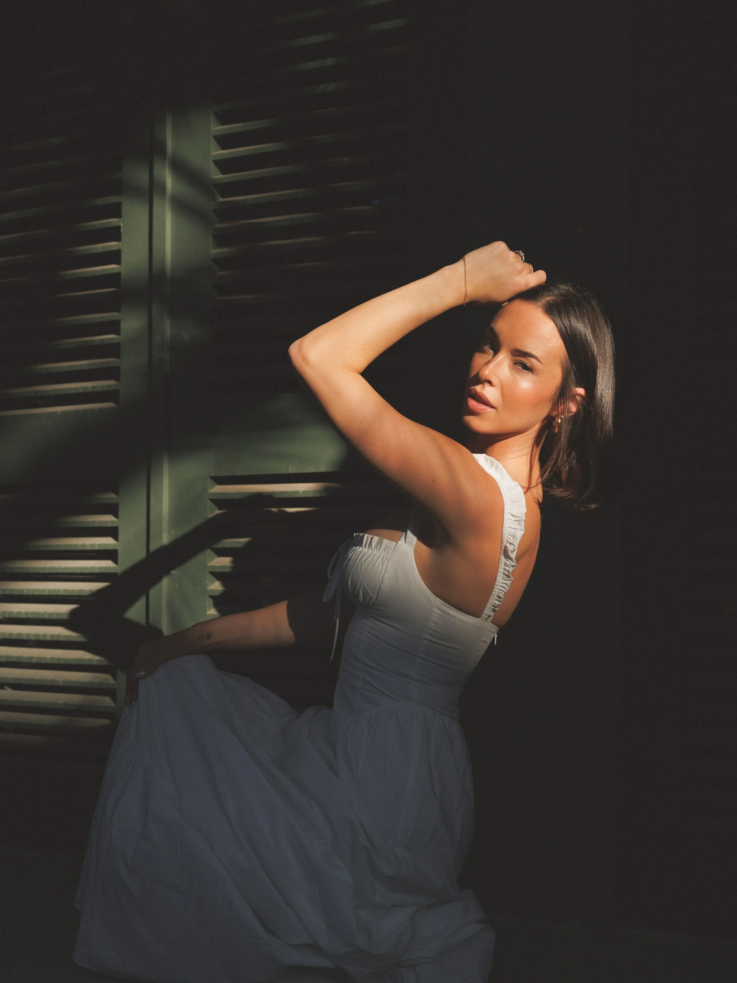 A woman in a white dress posing with her hand on her head, standing in front of green window shutters with shadows cast across her face and body.