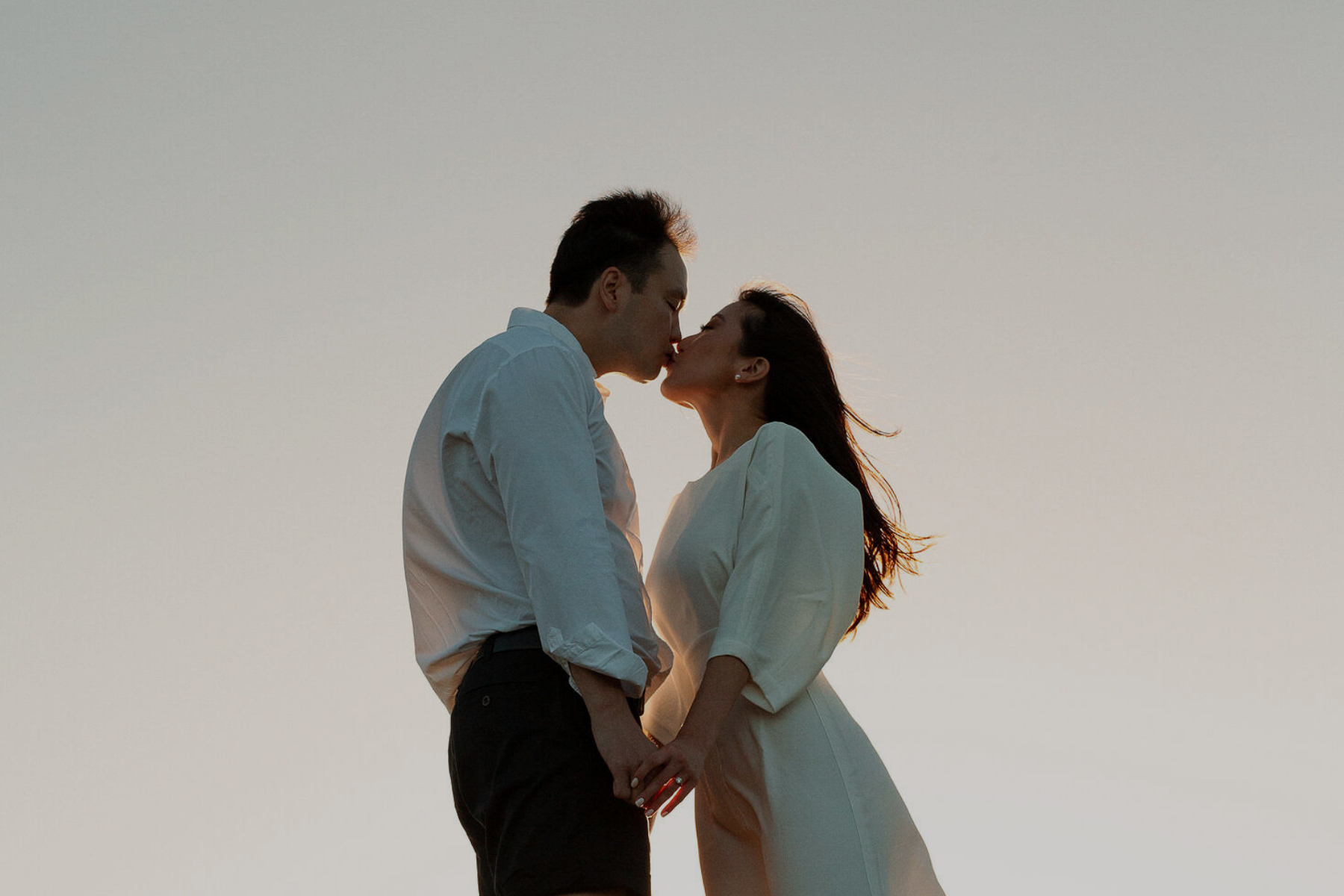 A couple kissing during sunset, holding hands, with a clear sky background.