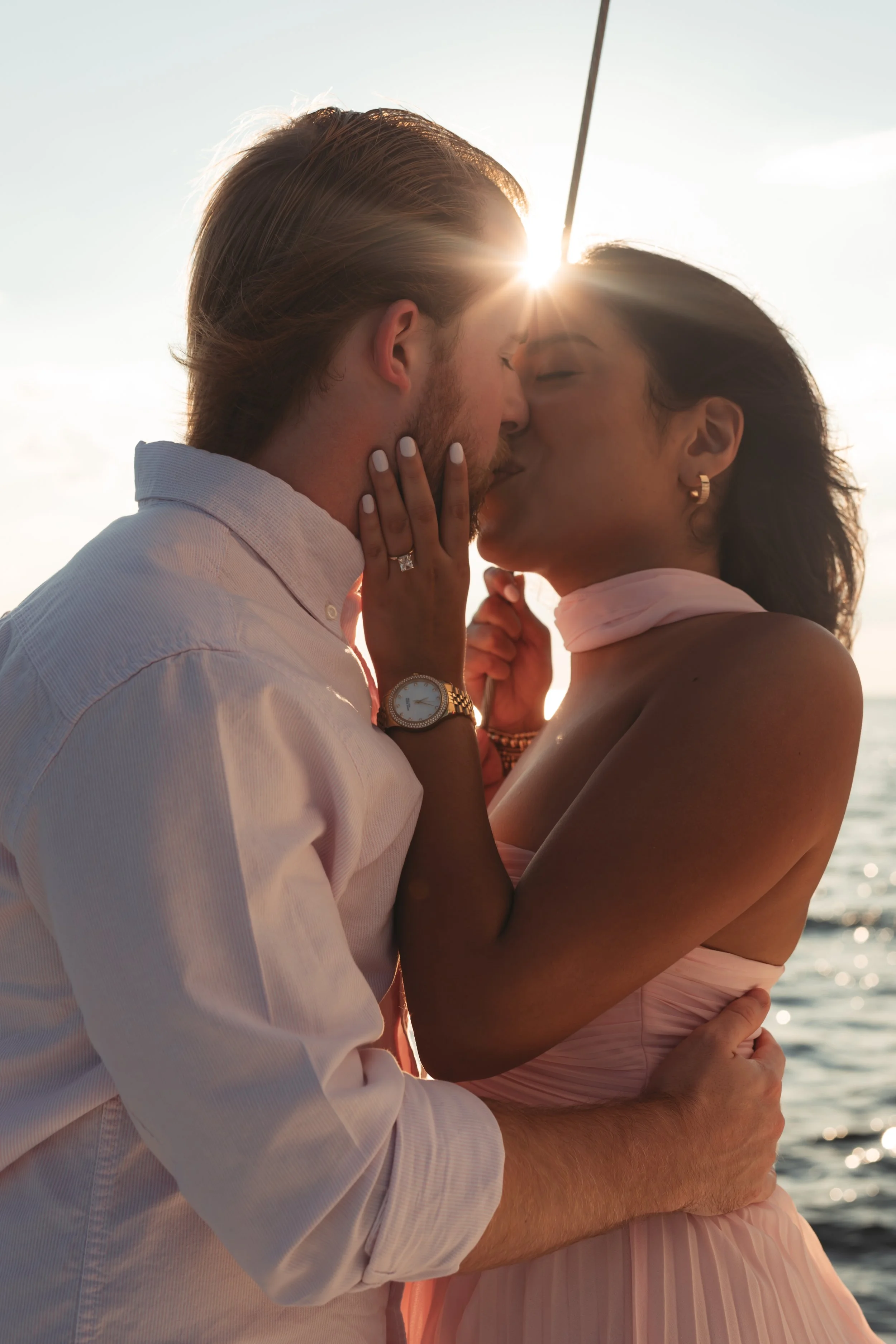 newly engaged couple kissing on a beach at sunset, with the sun shining behind them.