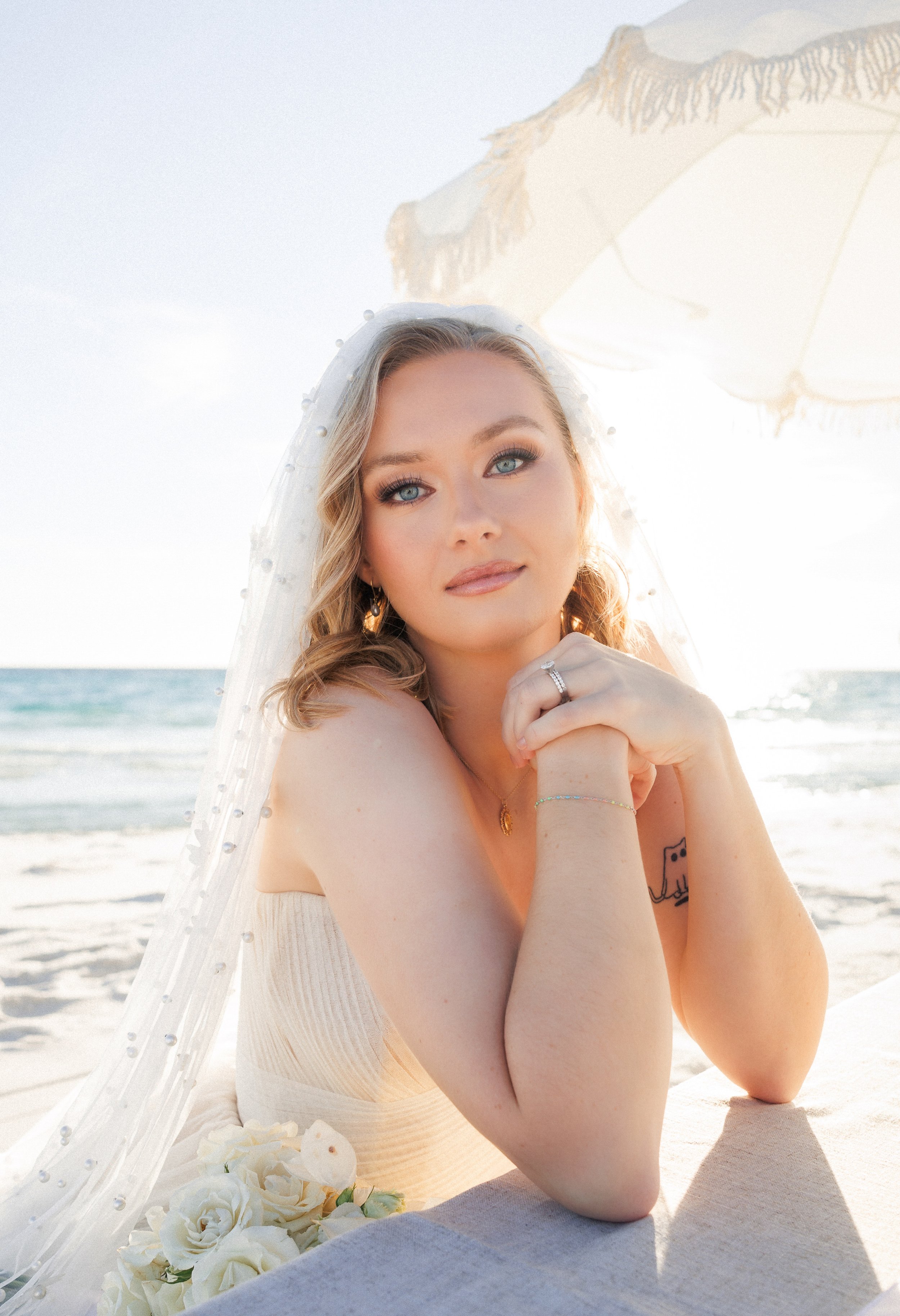 A woman sitting at the beach wearing a white sleeveless dress and a veil over her head, with the ocean and a large beach umbrella in the background.
