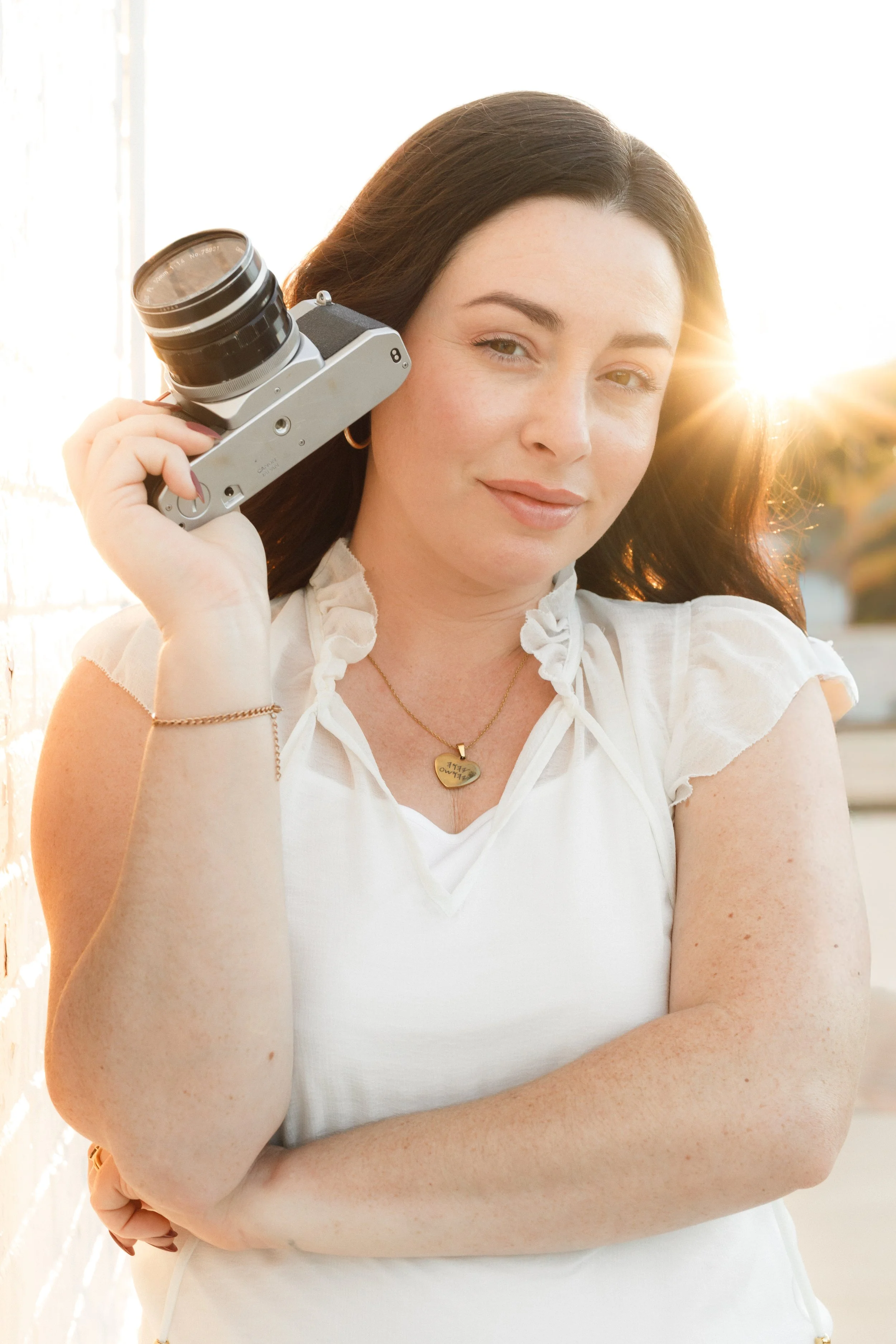 A woman with dark hair holding a vintage camera to her ear, standing outdoors during sunset, wearing a white top and jewelry.
