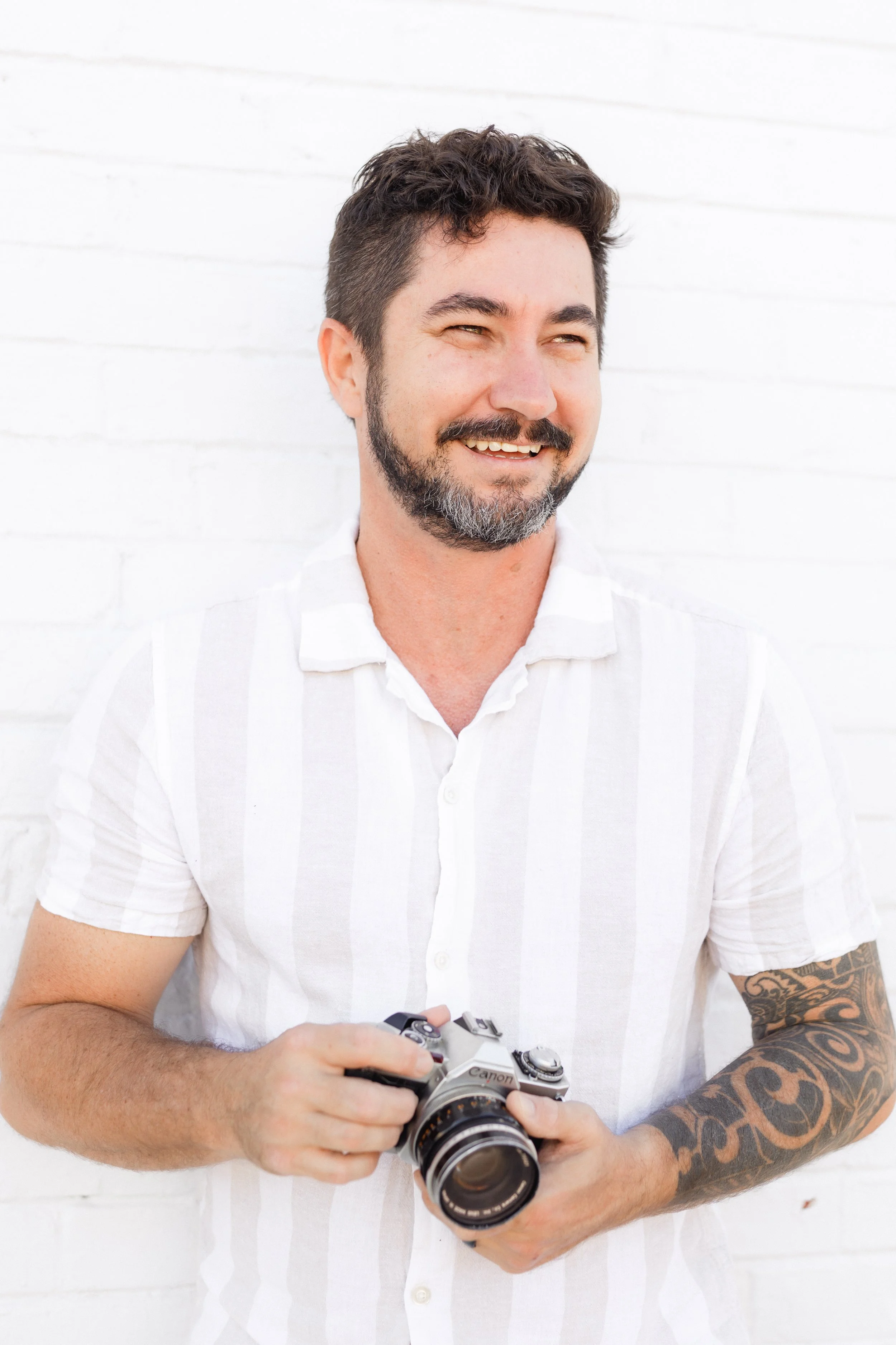 A man with dark hair, a beard, and tattoos on his right arm holding a camera, smiling, standing against a white brick wall.