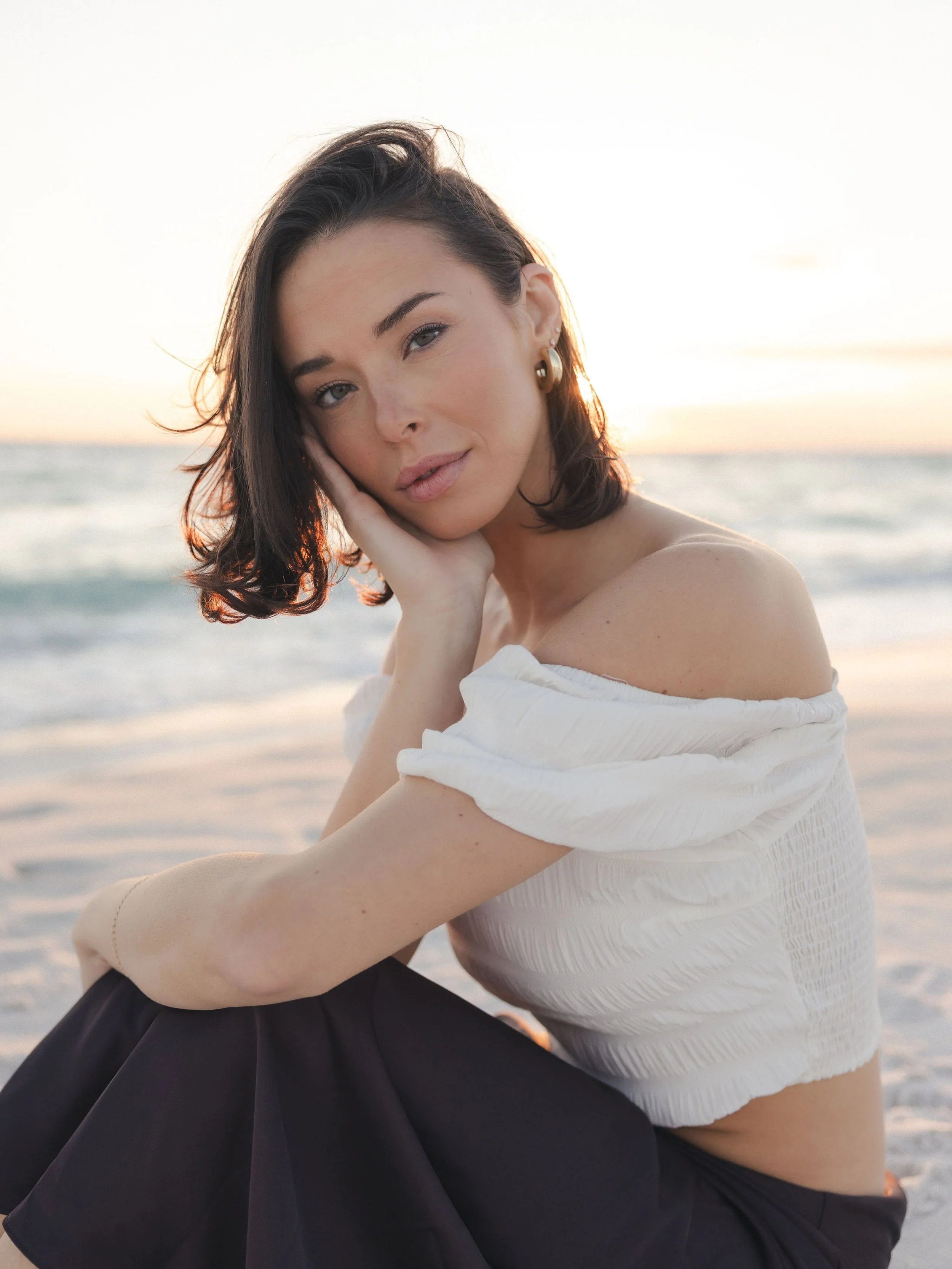 A woman with short dark hair and gold hoop earrings is sitting on the beach at sunset wearing an off-the-shoulder white top and black pants, looking at the camera with her hand resting on her cheek.