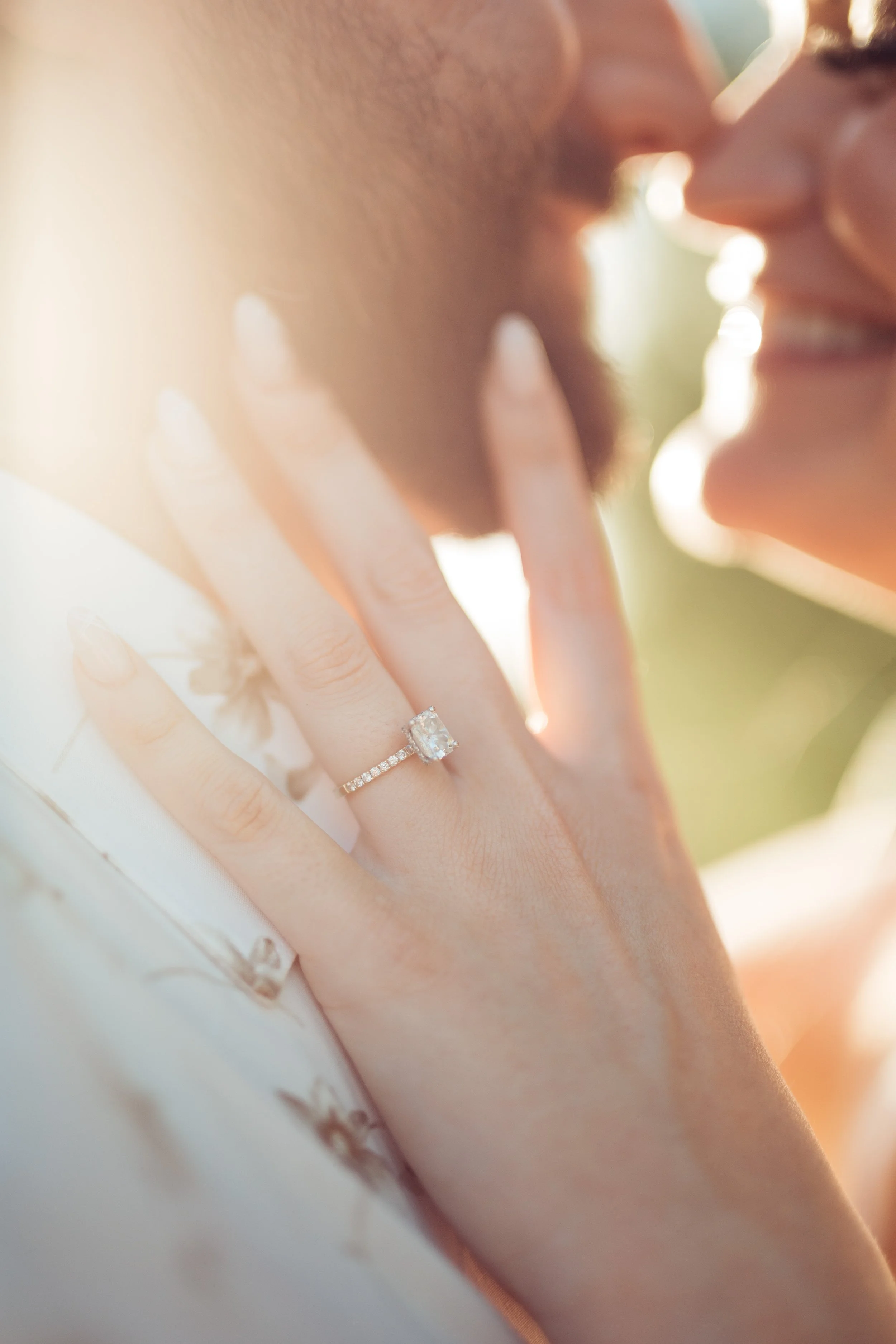 Close-up of a couple about to kiss, with focus on the woman's left hand showing an engagement ring. The woman has a smile and the man has a beard and mustache. Sunlight creates a warm glow.