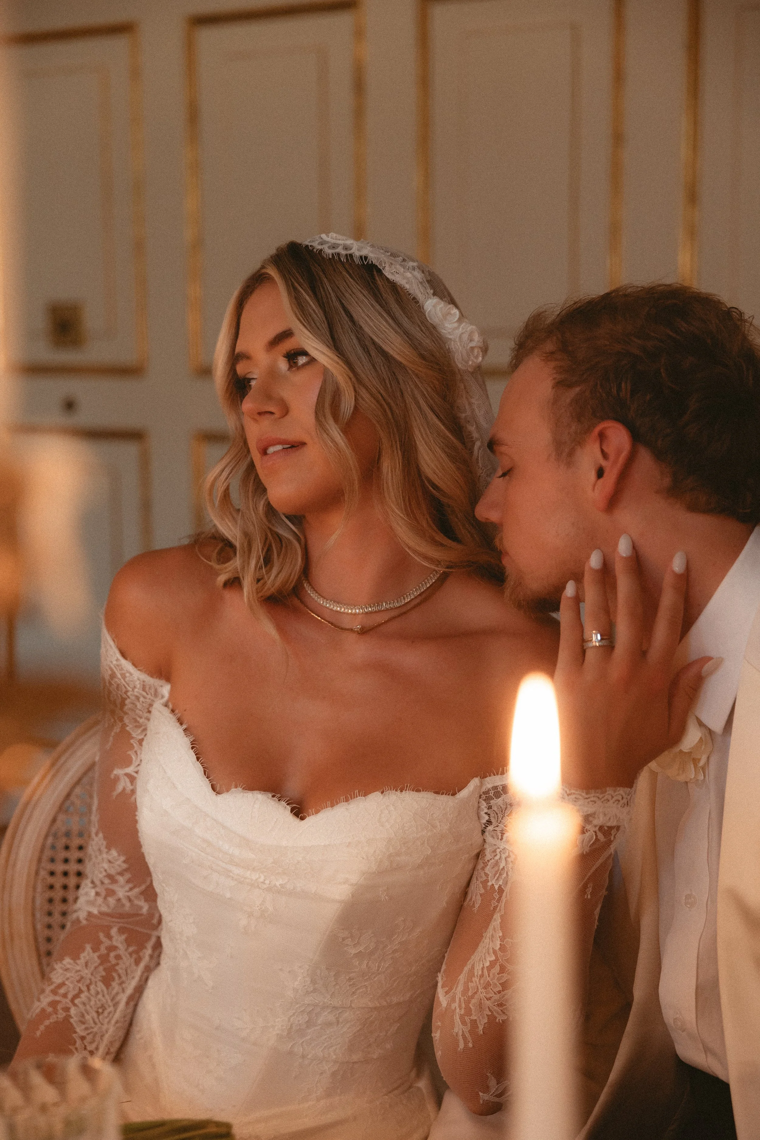 A bride in a lace wedding dress and a groom in a white suit share an intimate moment, with the groom gently resting his head near the bride's face, during a wedding celebration illuminated by candlelight.