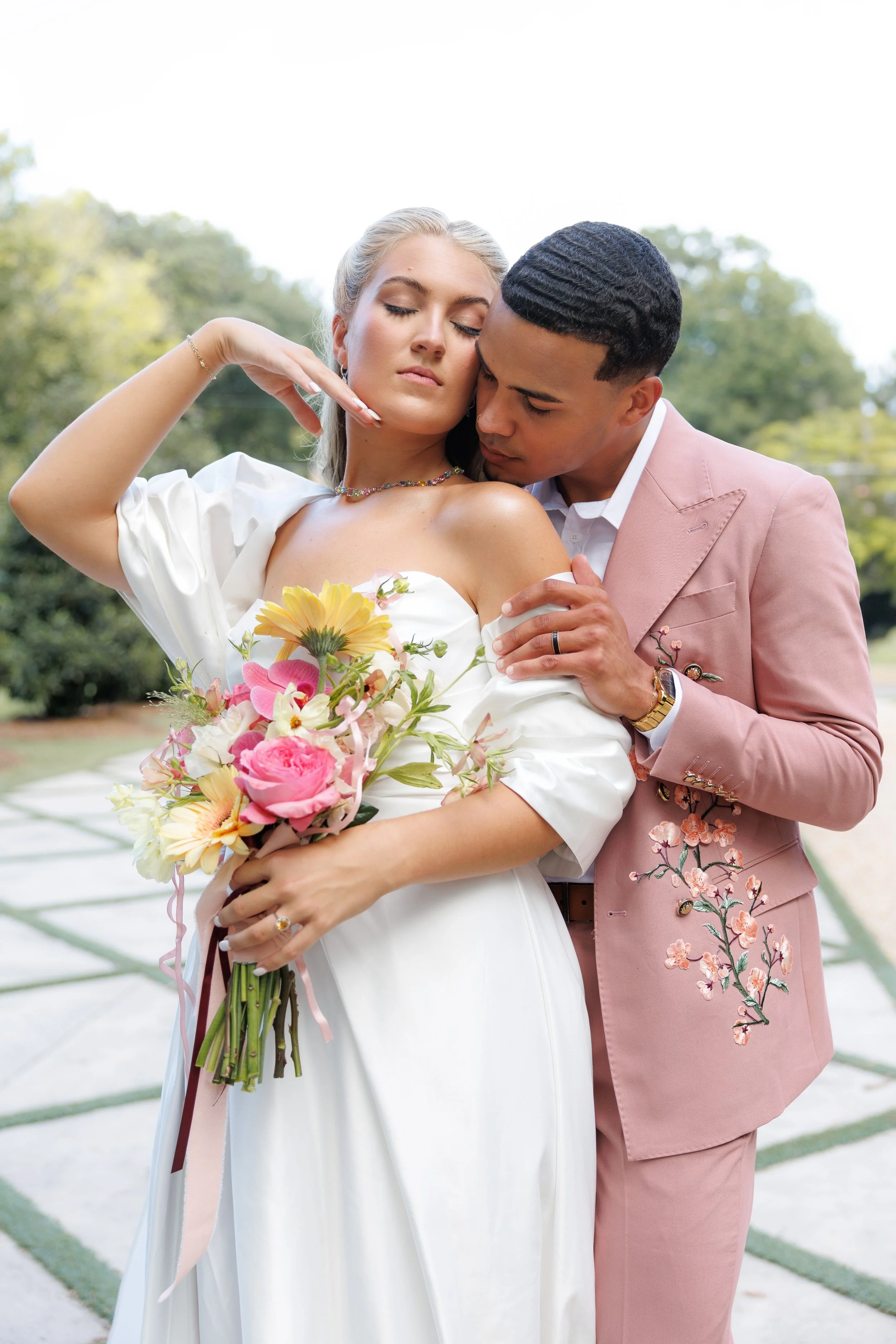 A bride and groom embrace outdoors, with the bride holding a bouquet of pink and yellow flowers, and the groom in a pink suit with floral embroidery.