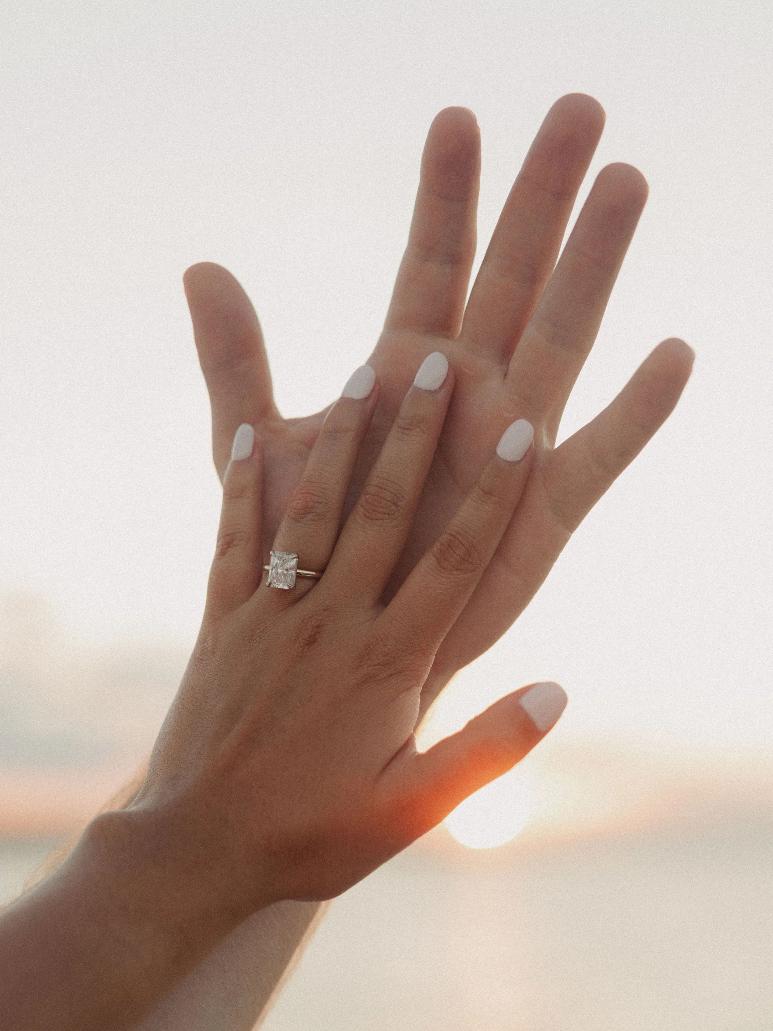 Two hands, one with a diamond ring, reaching towards the sunset over the water.