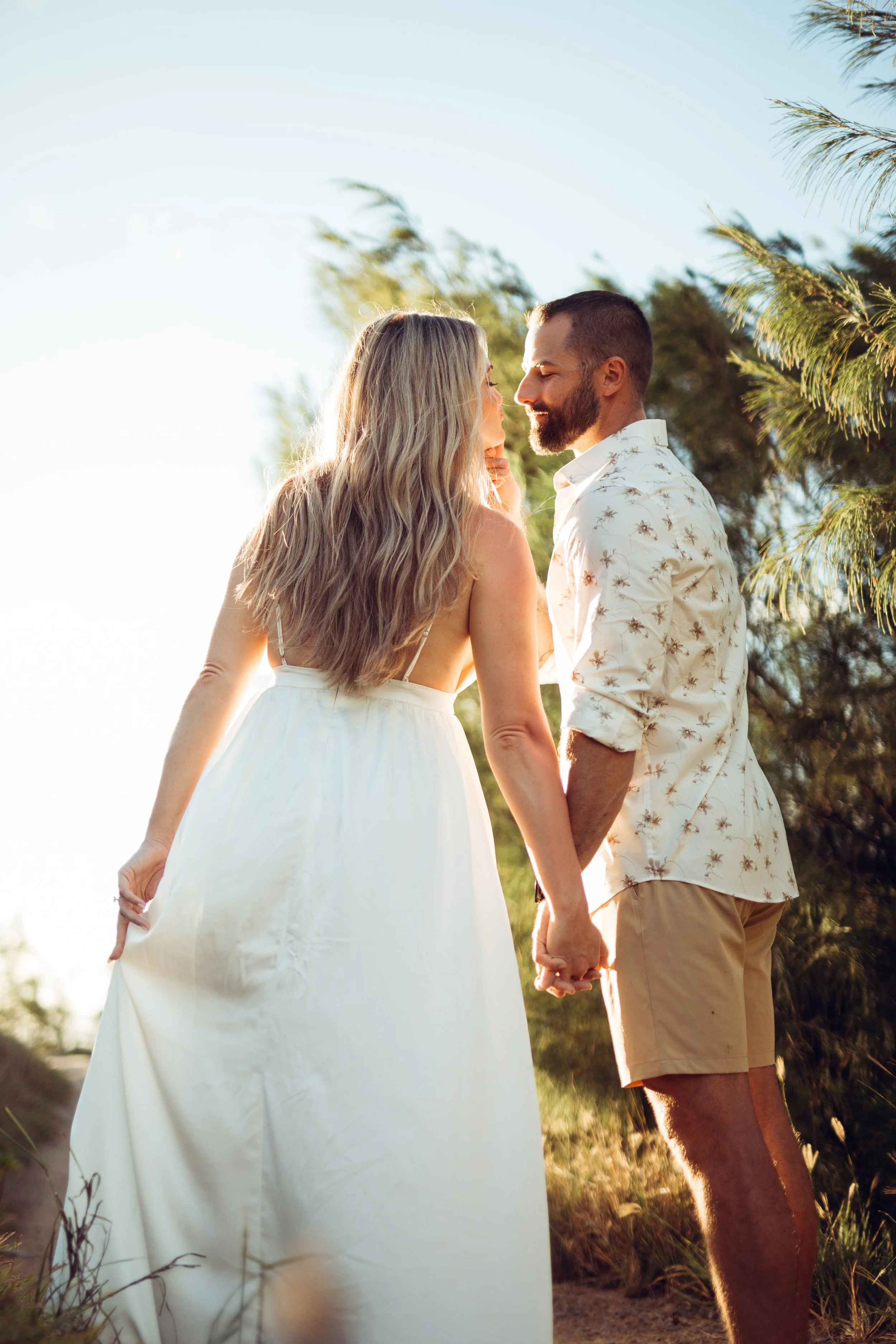 A couple holding hands and standing close together outdoors, facing each other in front of trees with sunlight shining behind them.