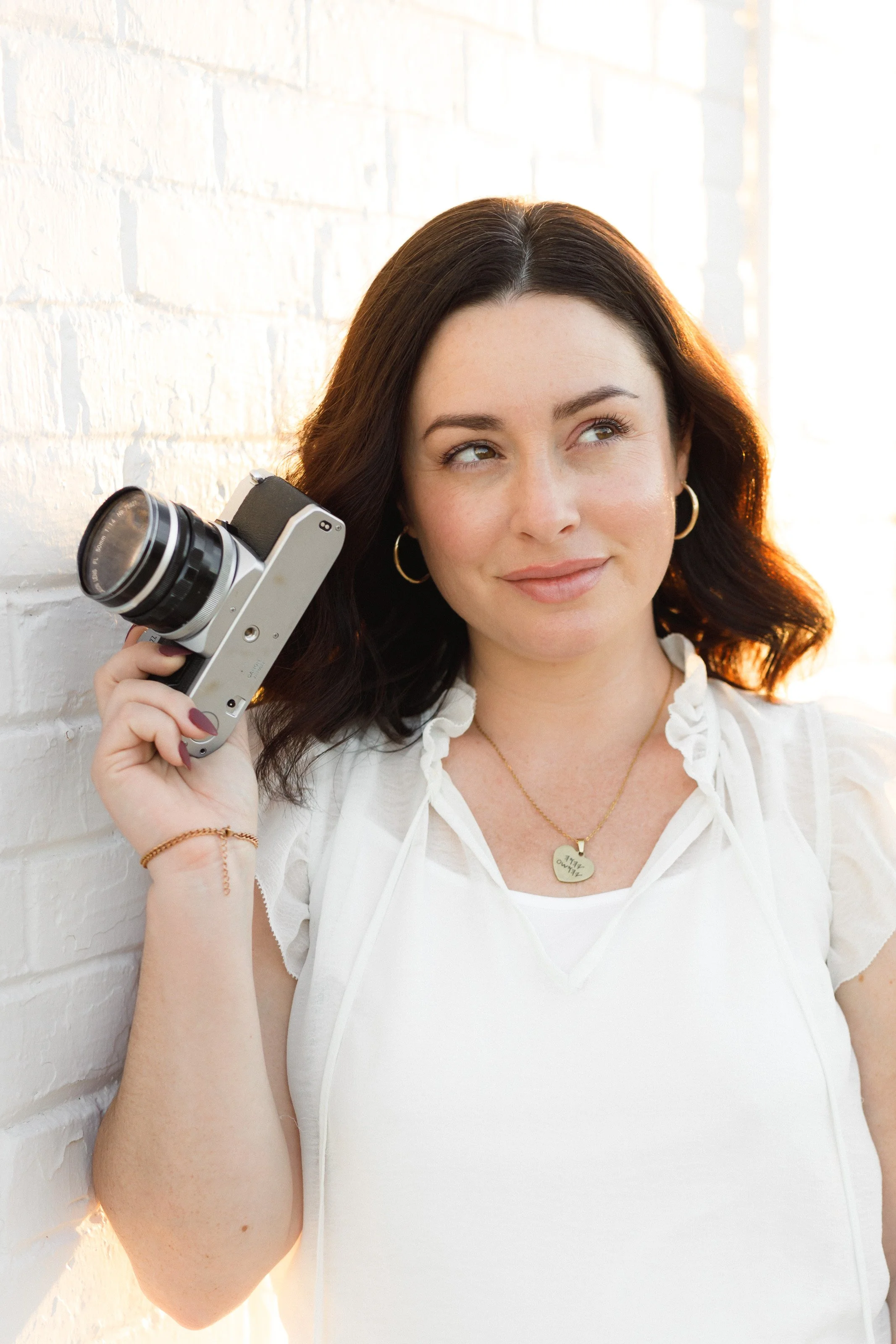 A woman with dark brown hair, wearing hoop earrings, a gold heart-shaped necklace, and a white top, is holding a vintage camera to her head and smiling thoughtfully while standing against a white brick wall with sunlight in the background.