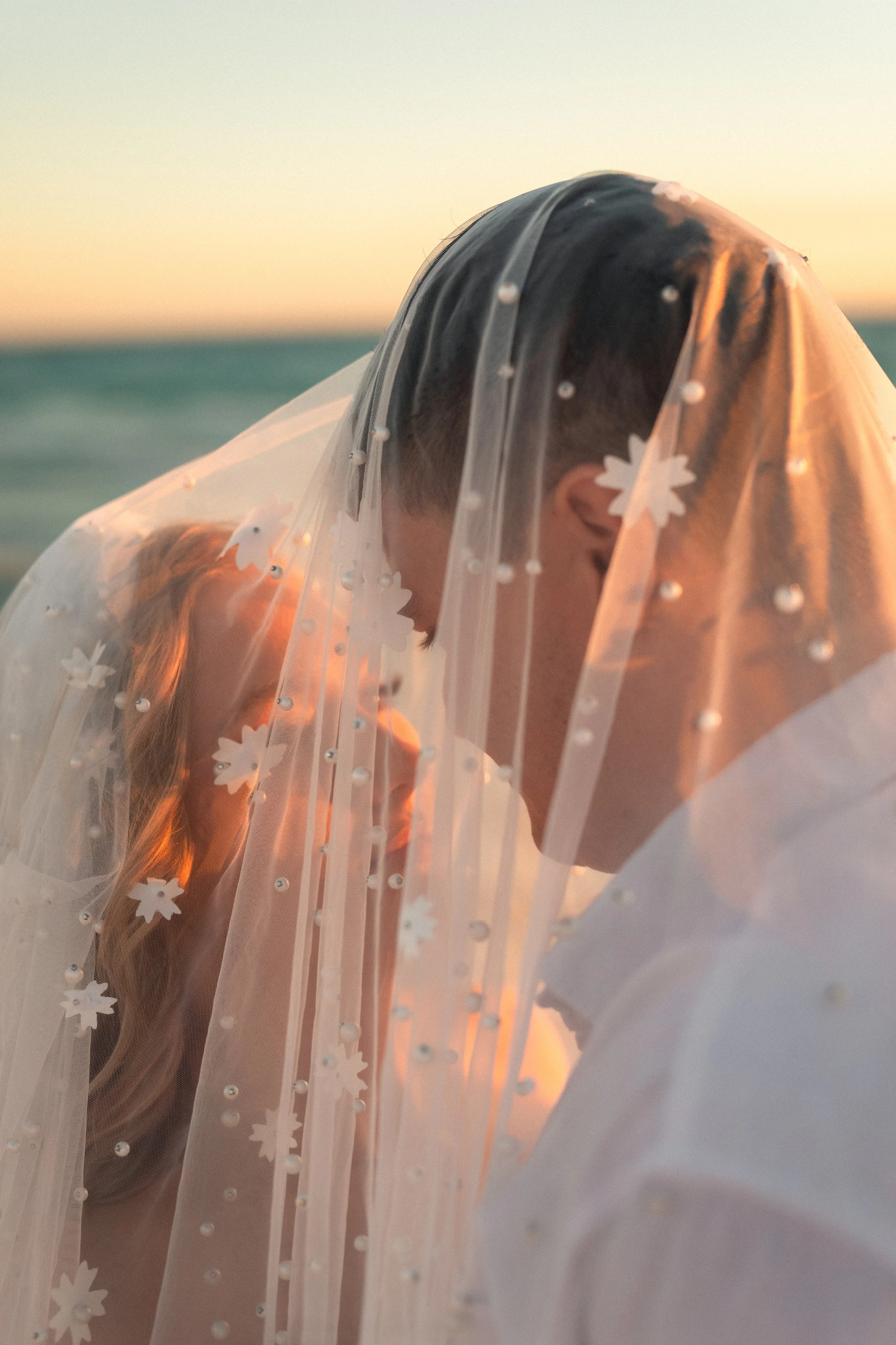 A couple on a beach under a sunset, with the bridal veil covering their faces as they nuzzle close.