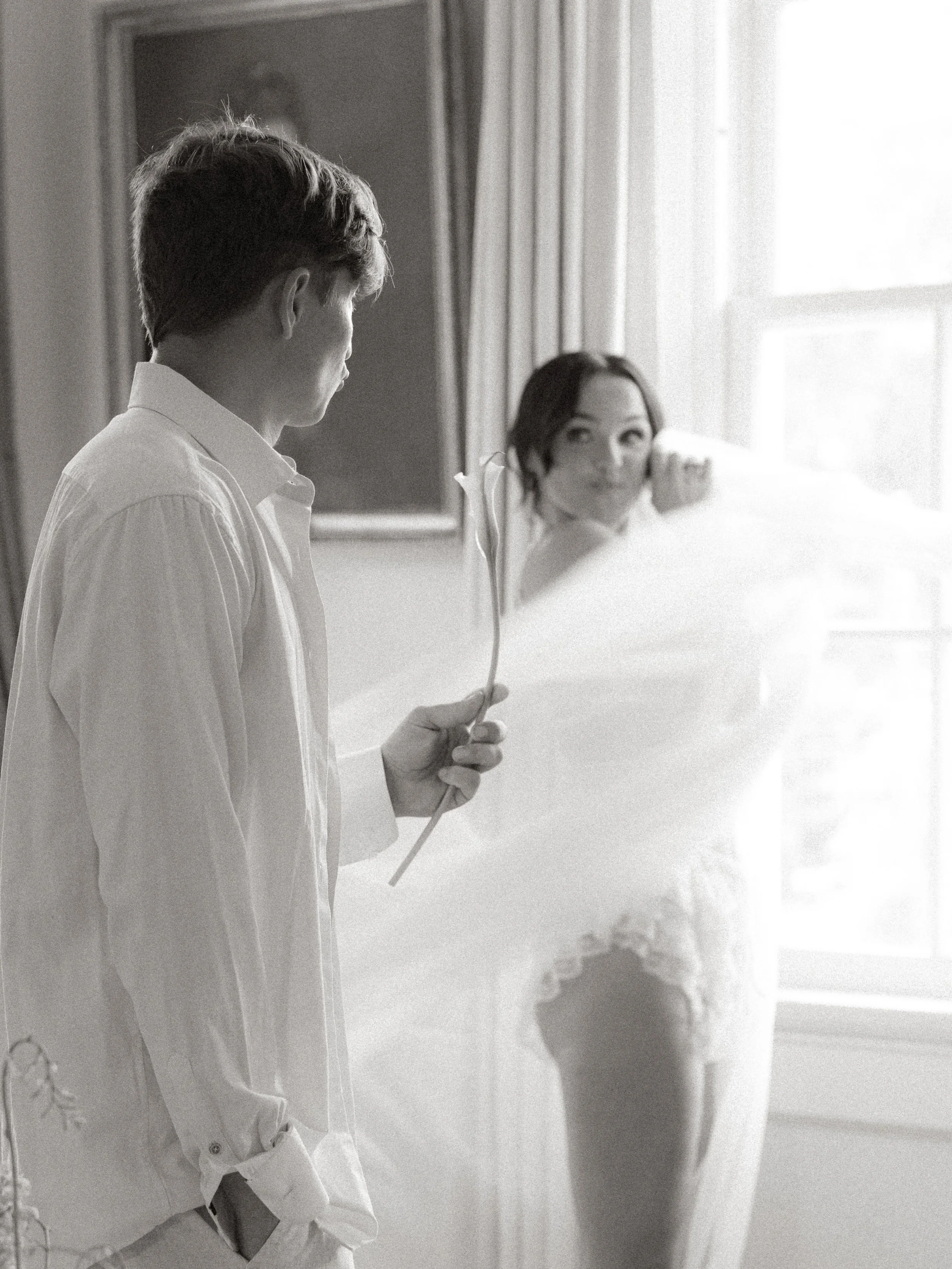 A man holding a single flower and a woman in a wedding dress looking at him in a room with a window and curtains.