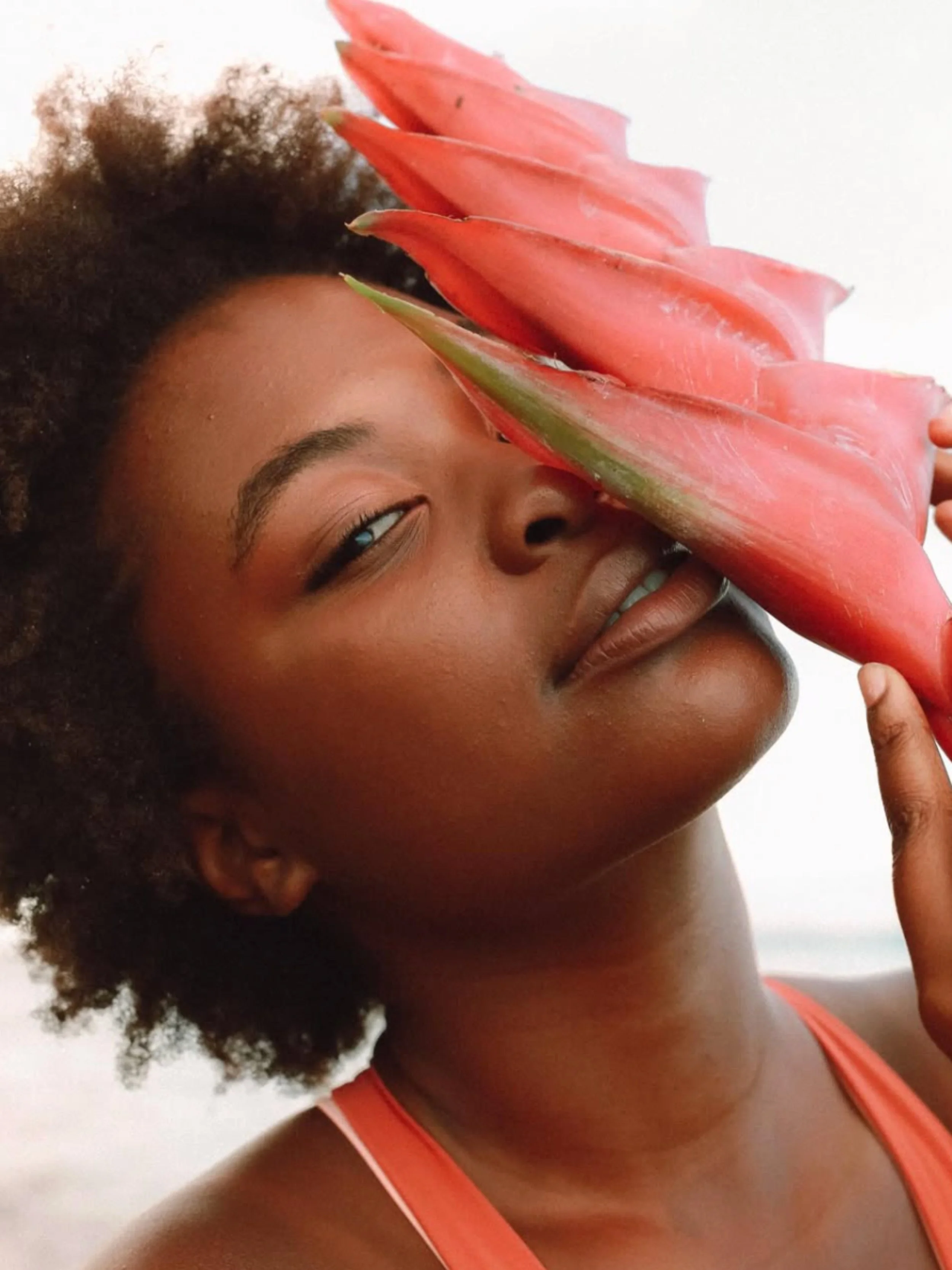 A woman with curly hair holding a large pink tropical flower close to her face, partially covering her eye, with a serene expression.