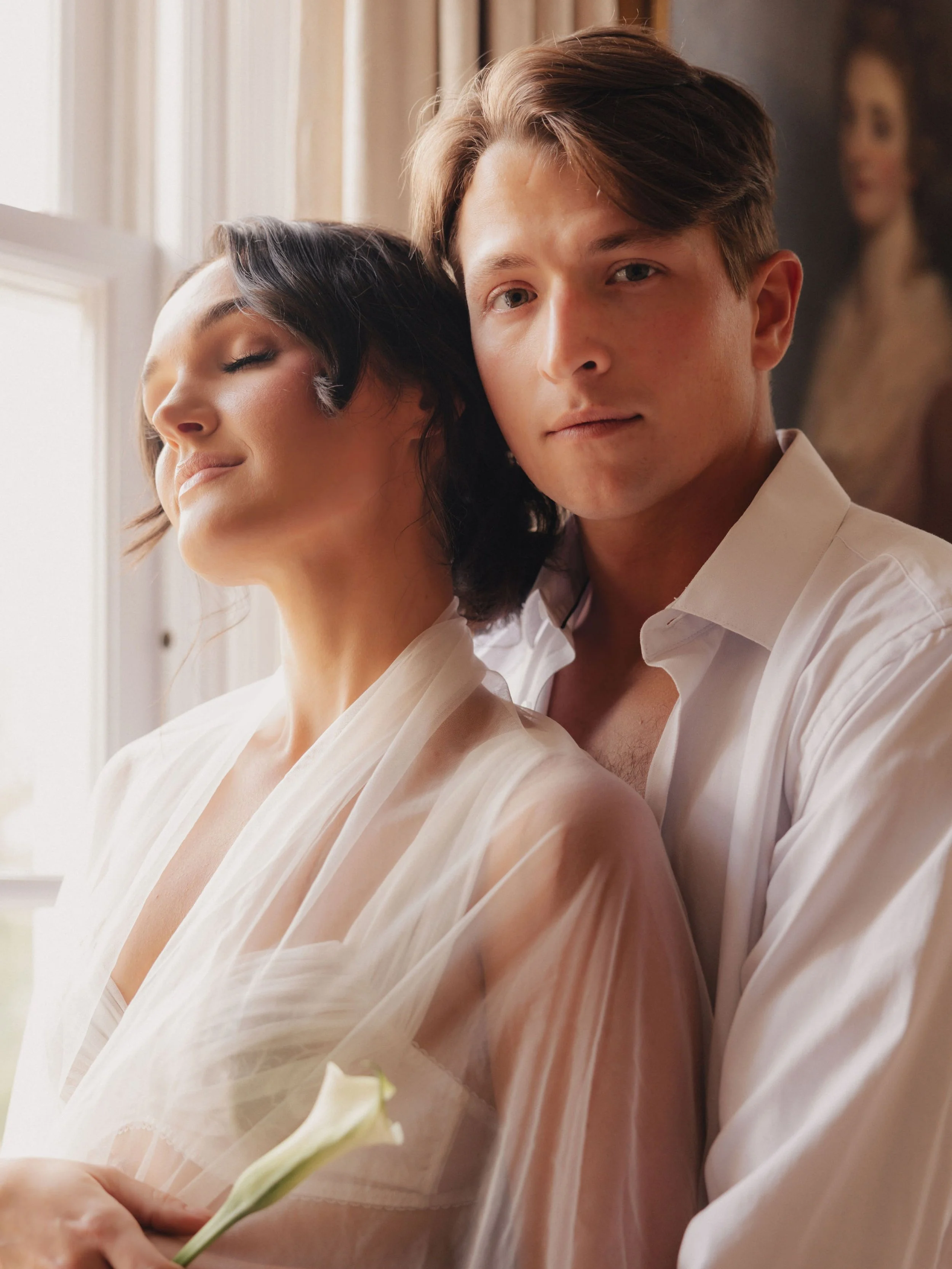 A man and woman in an intimate pose indoors with natural light, woman with eyes closed and holding a white flower, man looking at the camera, both dressed in white