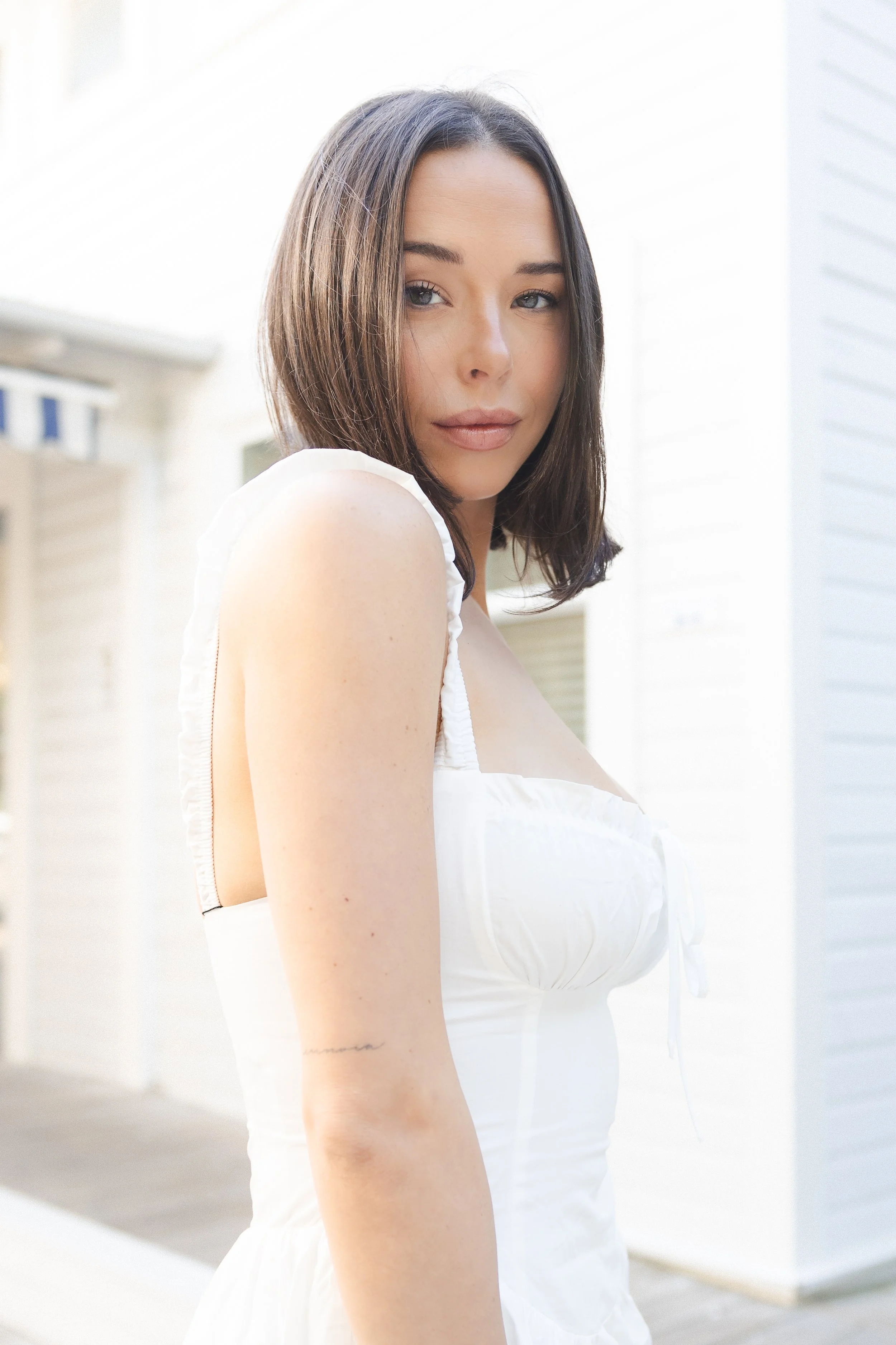 A woman with dark brown hair and fair skin is looking at the camera while wearing a white sleeveless dress. She is outdoors with a background of white siding and sunlight.