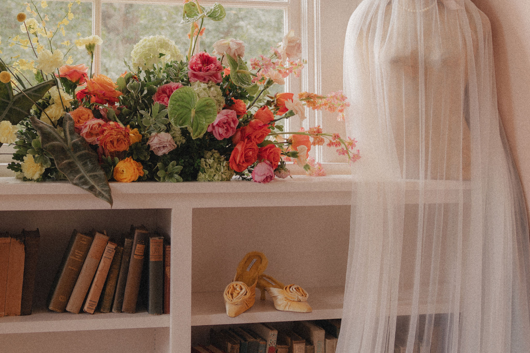 A white bookshelf with books and a pair of yellow satin high-heeled shoes on the middle shelf, and a large bouquet of colorful flowers on top, near a window with sheer curtains.