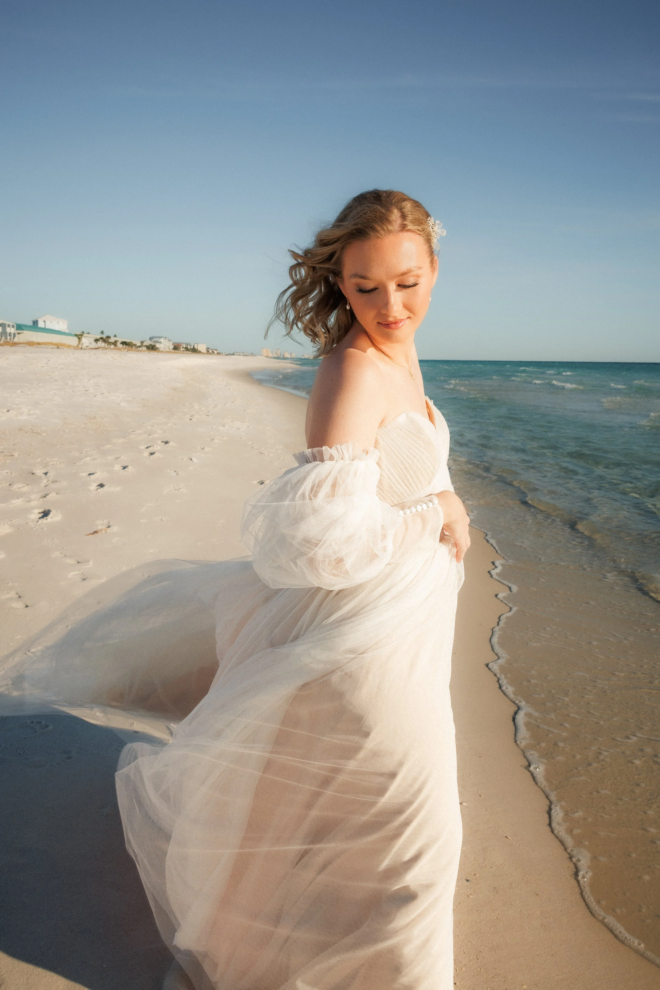 A woman in a wedding dress standing on the beach with waves and a clear sky in the background.