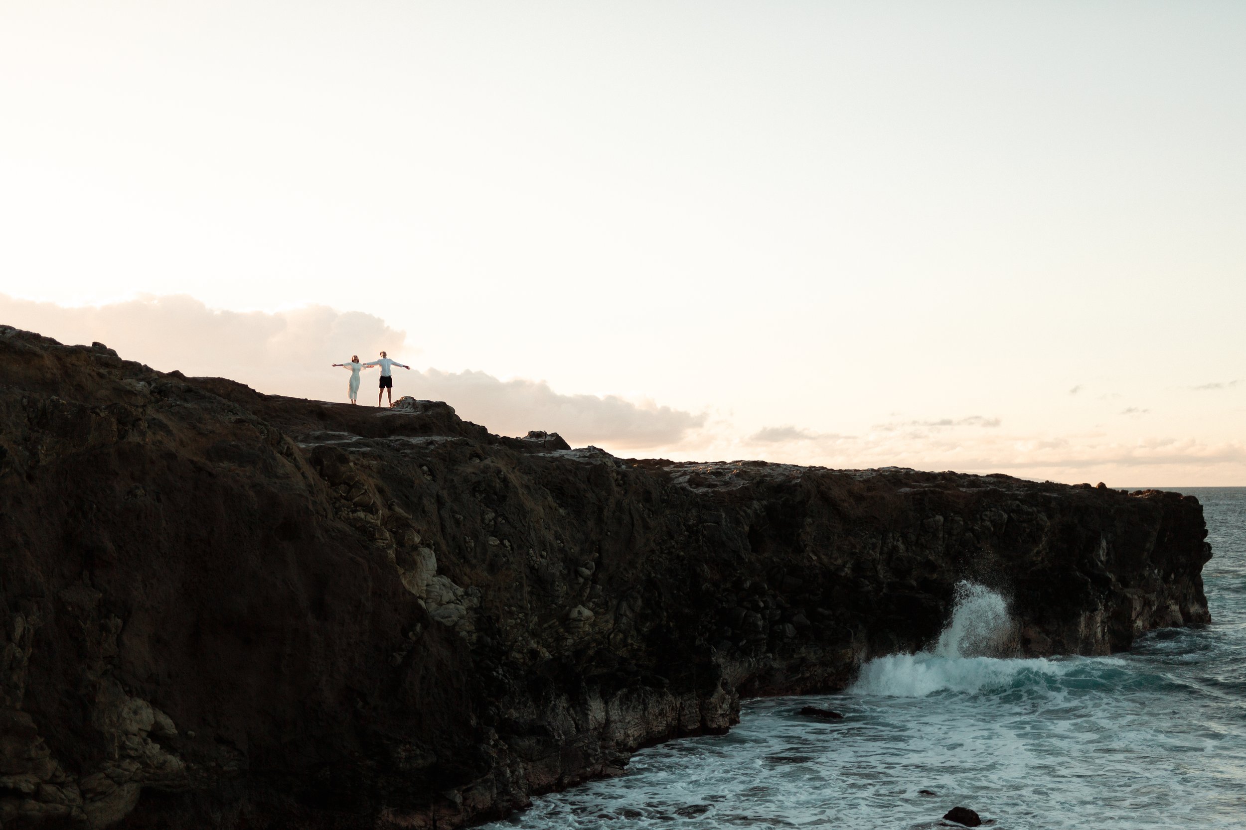 Two people stand on a rocky cliff by the ocean with arms outstretched as the sun sets or rises in the background