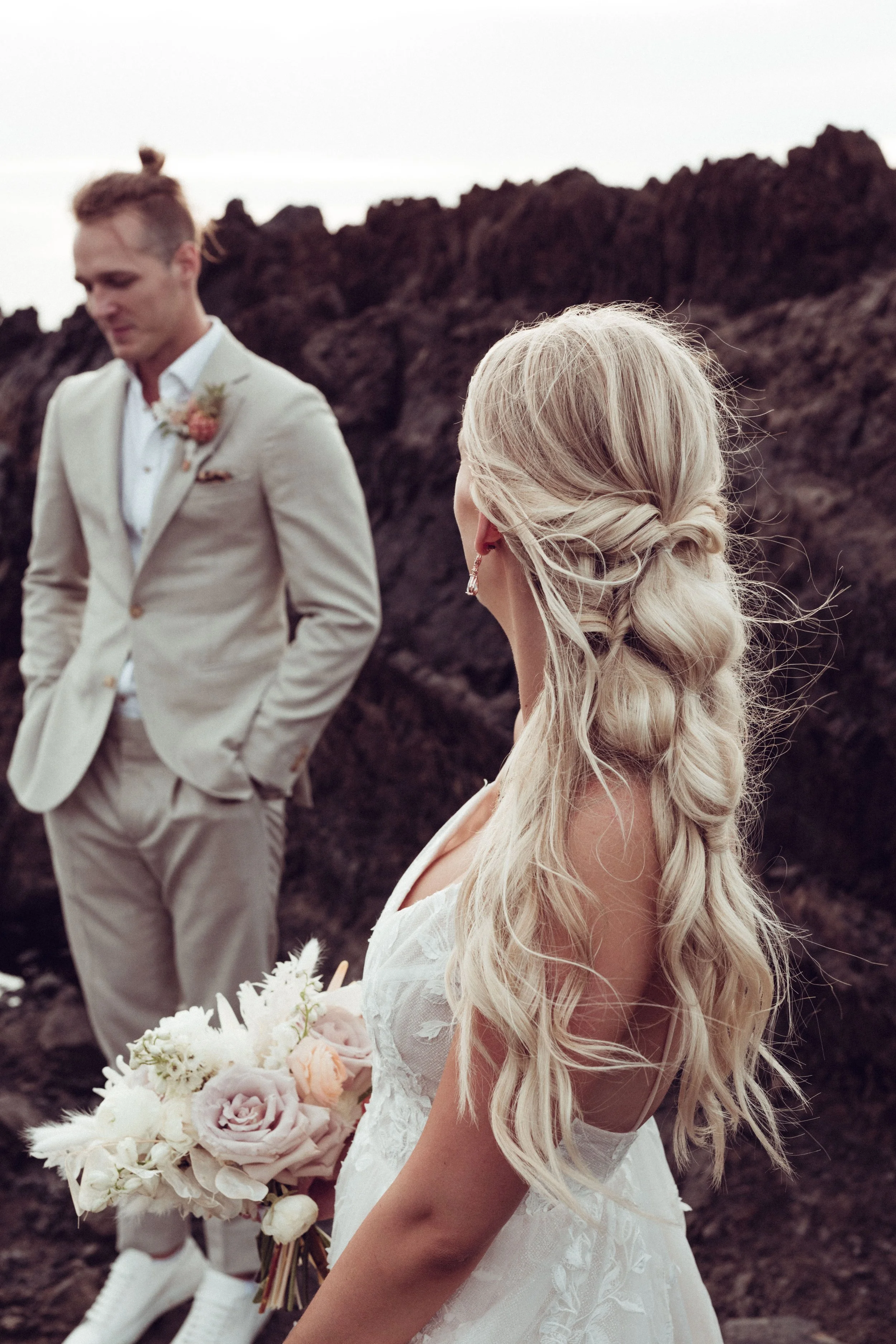 A bride in a white dress holding a bouquet of pink and white roses with a groom in a beige suit behind her, set against a rocky outdoor backdrop.