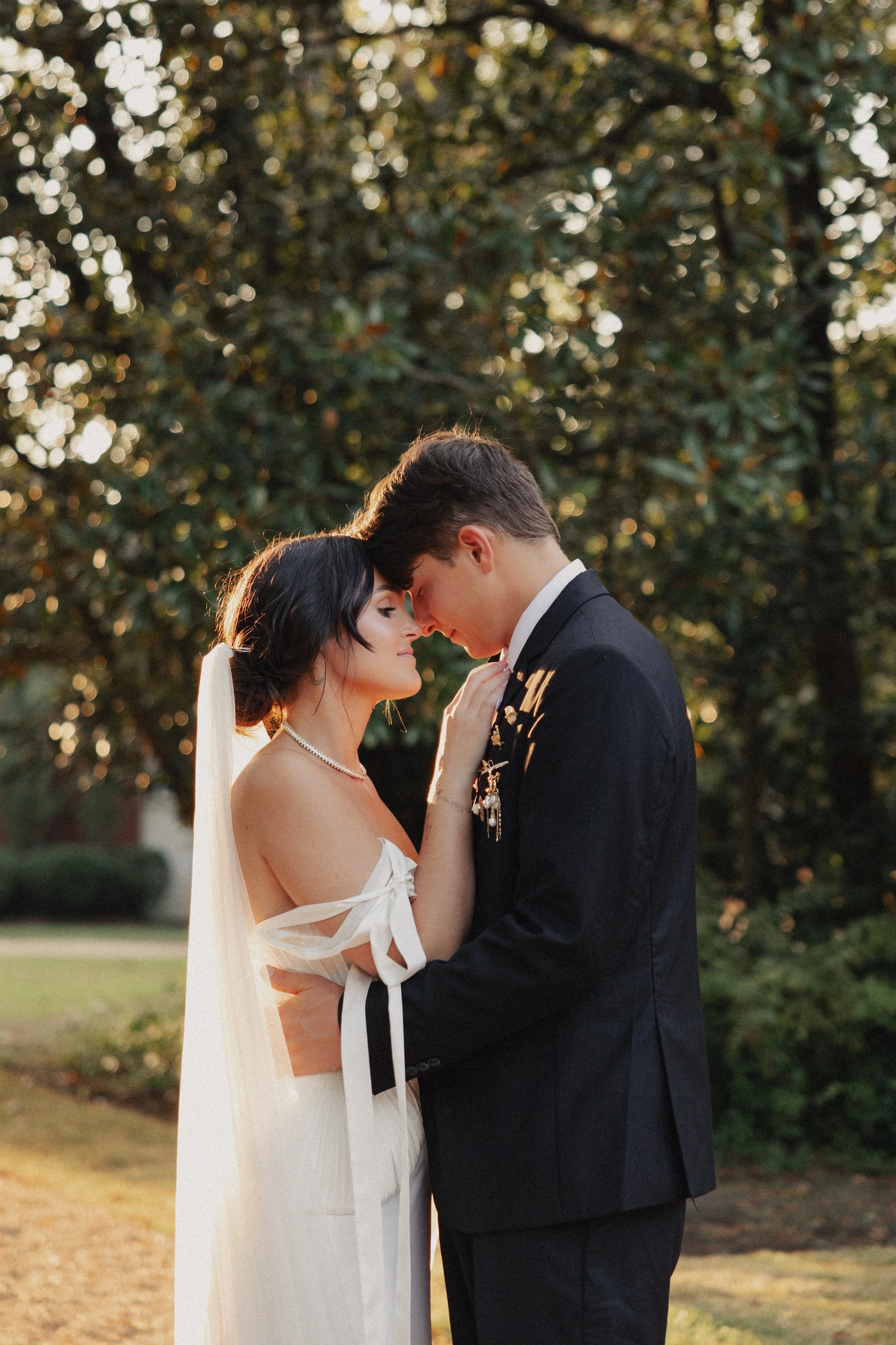 A bride and groom embrace outdoors at sunset, with the bride wearing a white off-shoulder dress and the groom in a black suit, their foreheads touching and eyes closed.