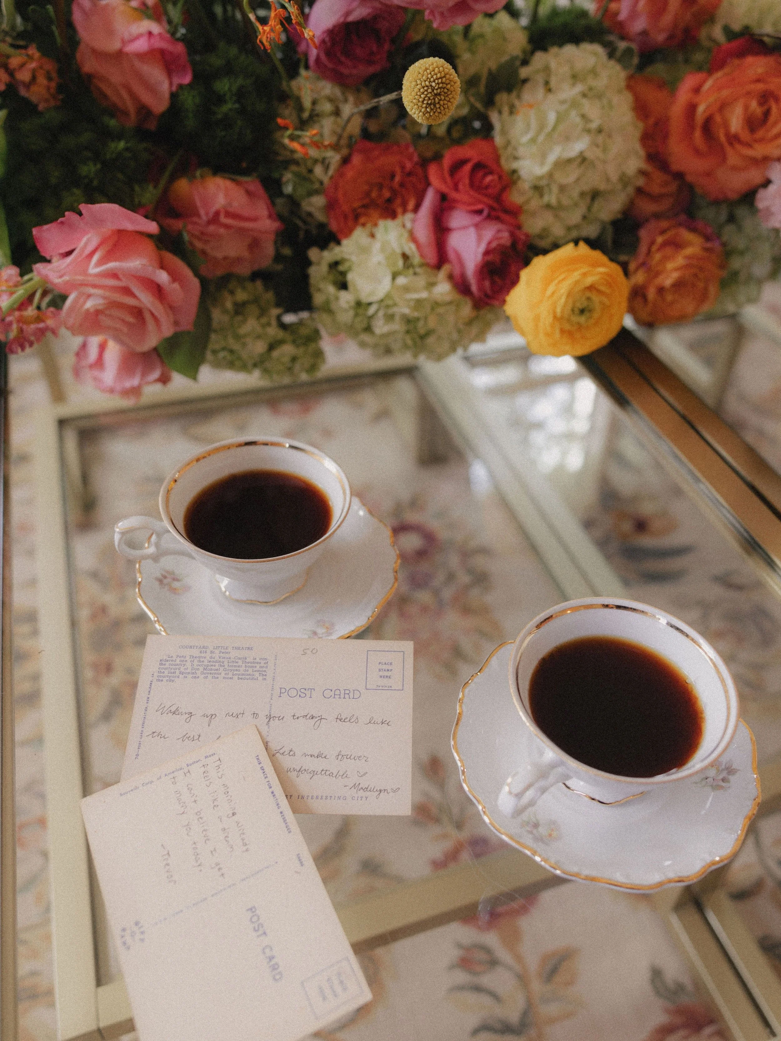 Two cups of black coffee in white porcelain cups with gold trim on a glass table, alongside a note and a floral arrangement with pink, peach, white, and yellow flowers.