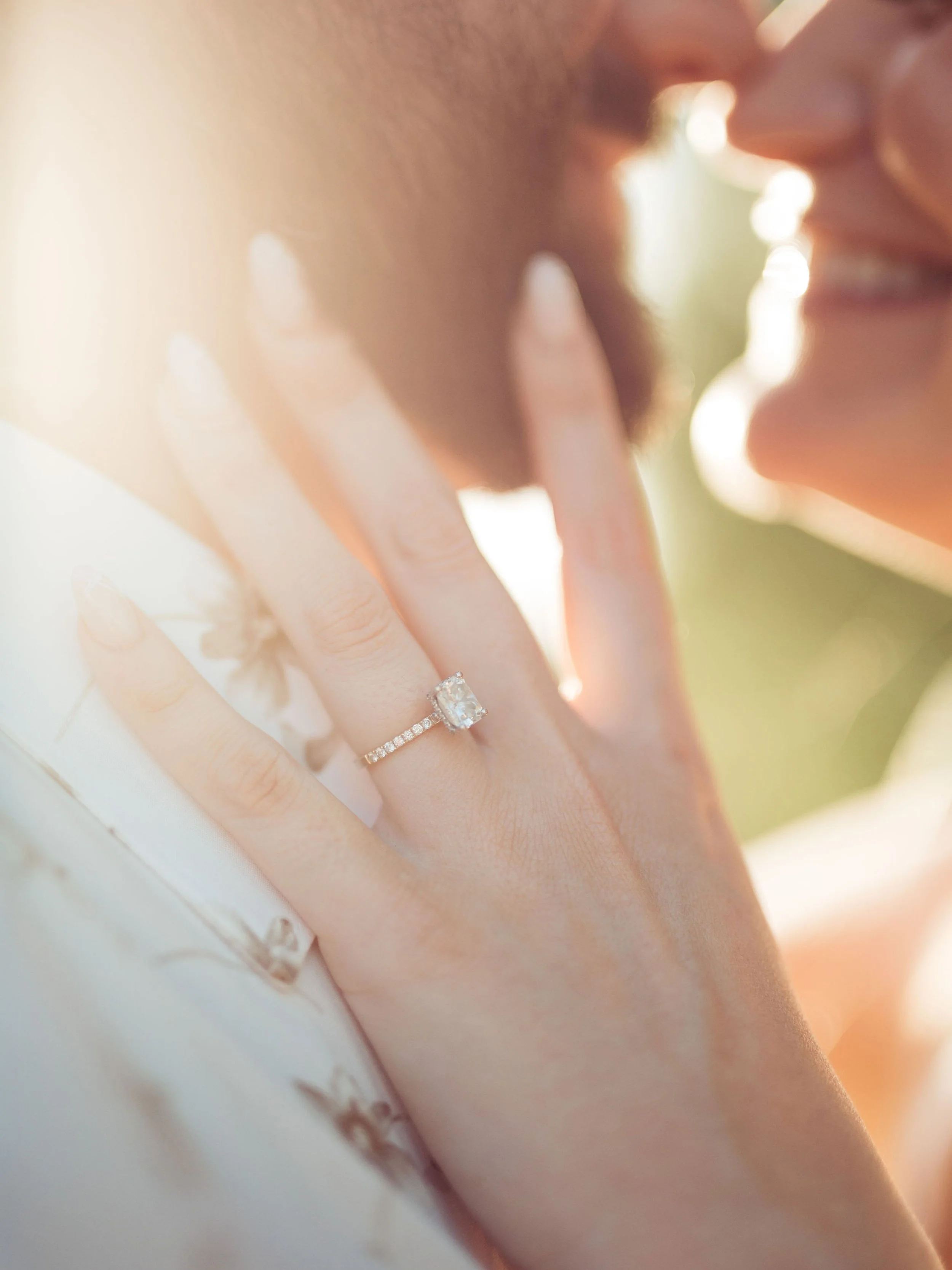Close-up of a woman's hand showing an engagement ring with a large square-cut diamond and a wedding band with smaller diamonds, with a blurred smiling couple in the background, touching noses.