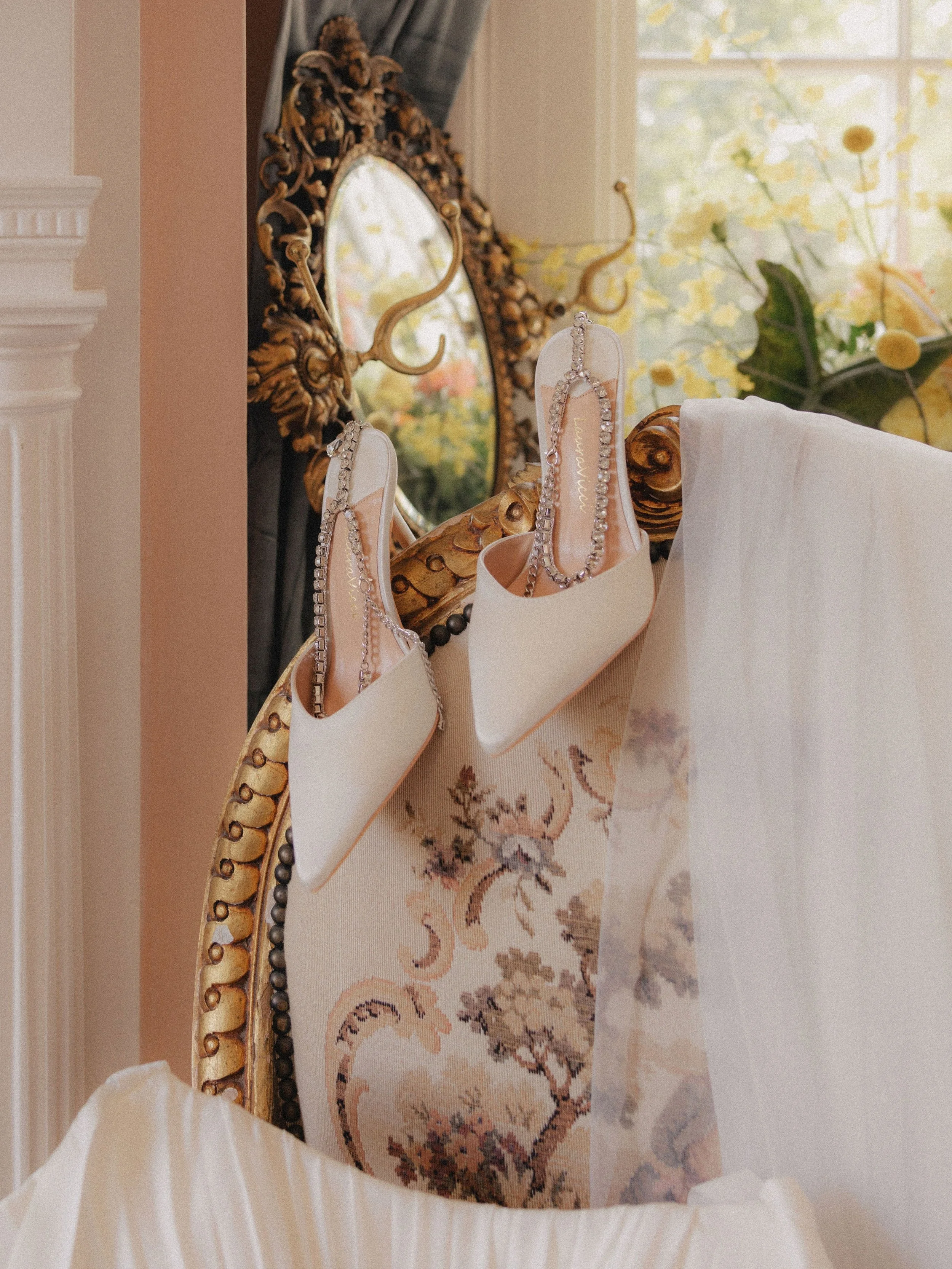 A pair of white high heel shoes with decorated straps draped over an ornate gold frame mirror, which is partially covered by draped white fabric, in a room decorated with floral patterns and a large window in the background.