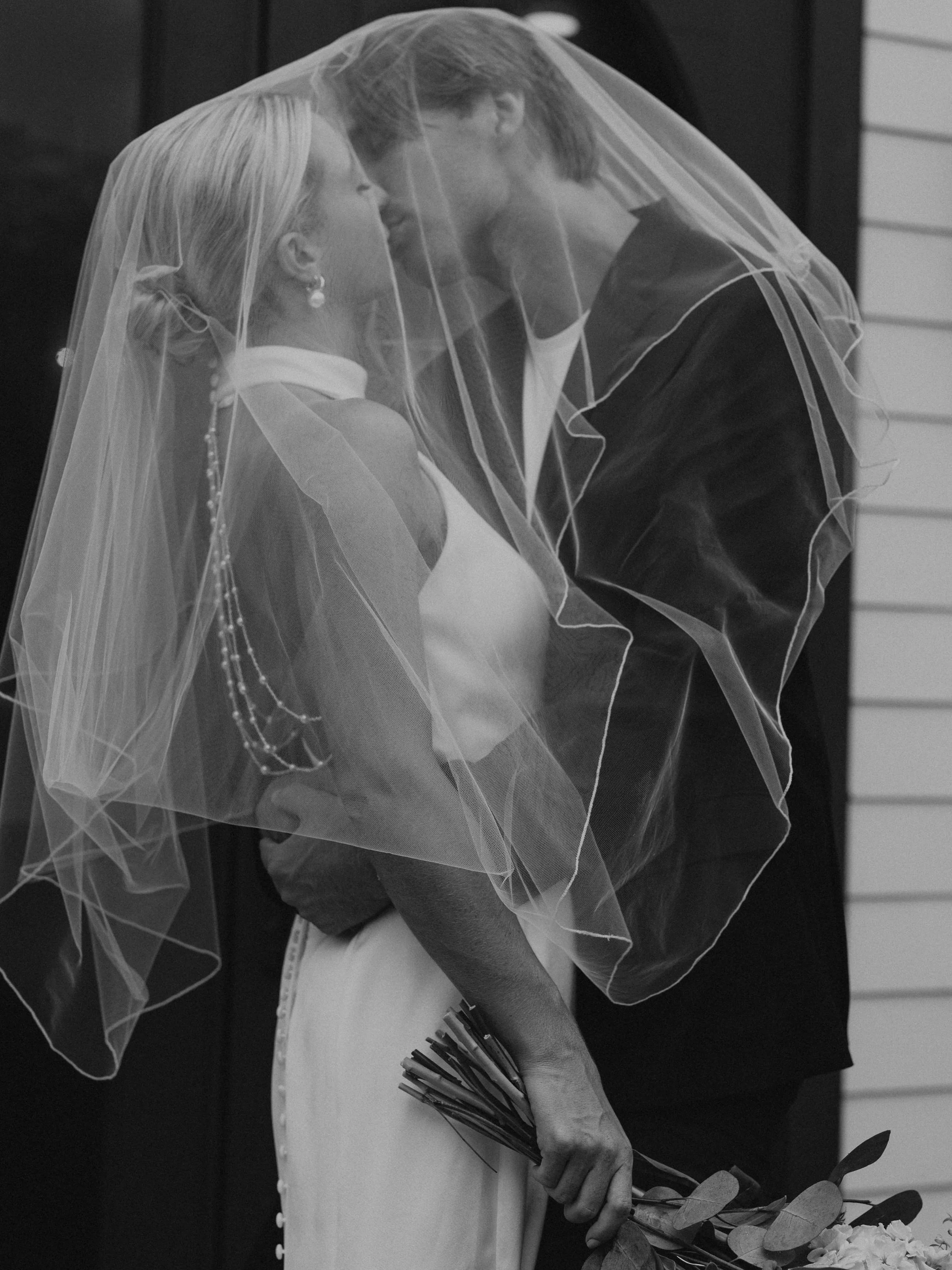 A bride and groom kissing under a wedding veil, with the bride holding a bouquet of flowers.