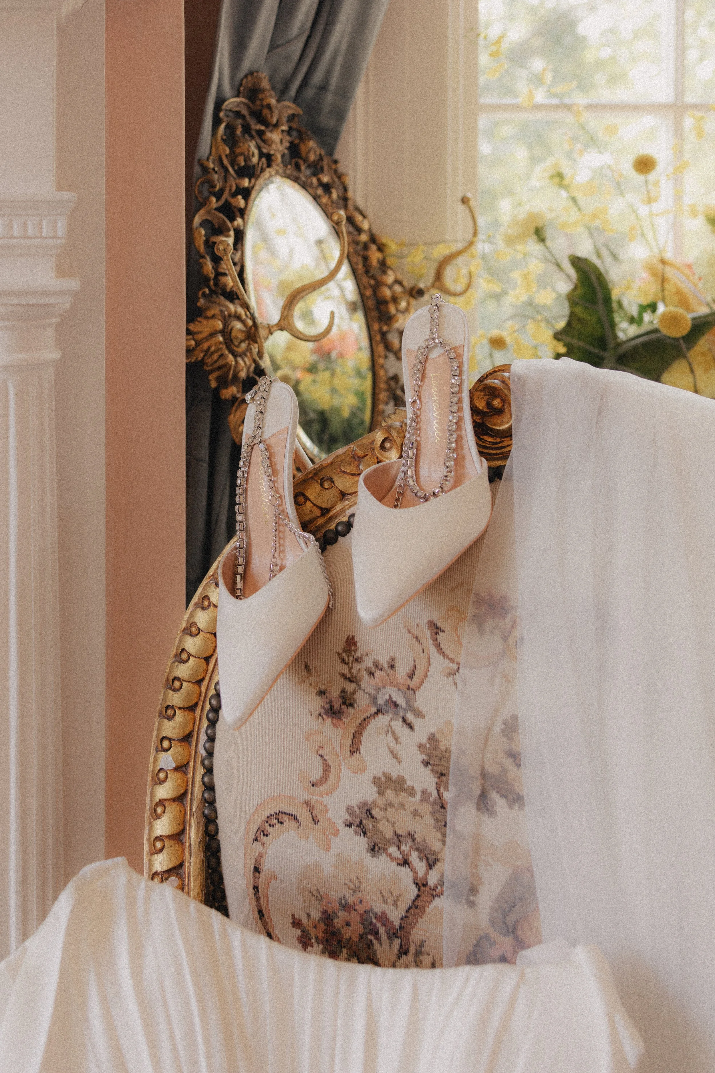 A pair of white high-heeled shoes with silver chains on a decorative chair, with a mirror and window in the background.