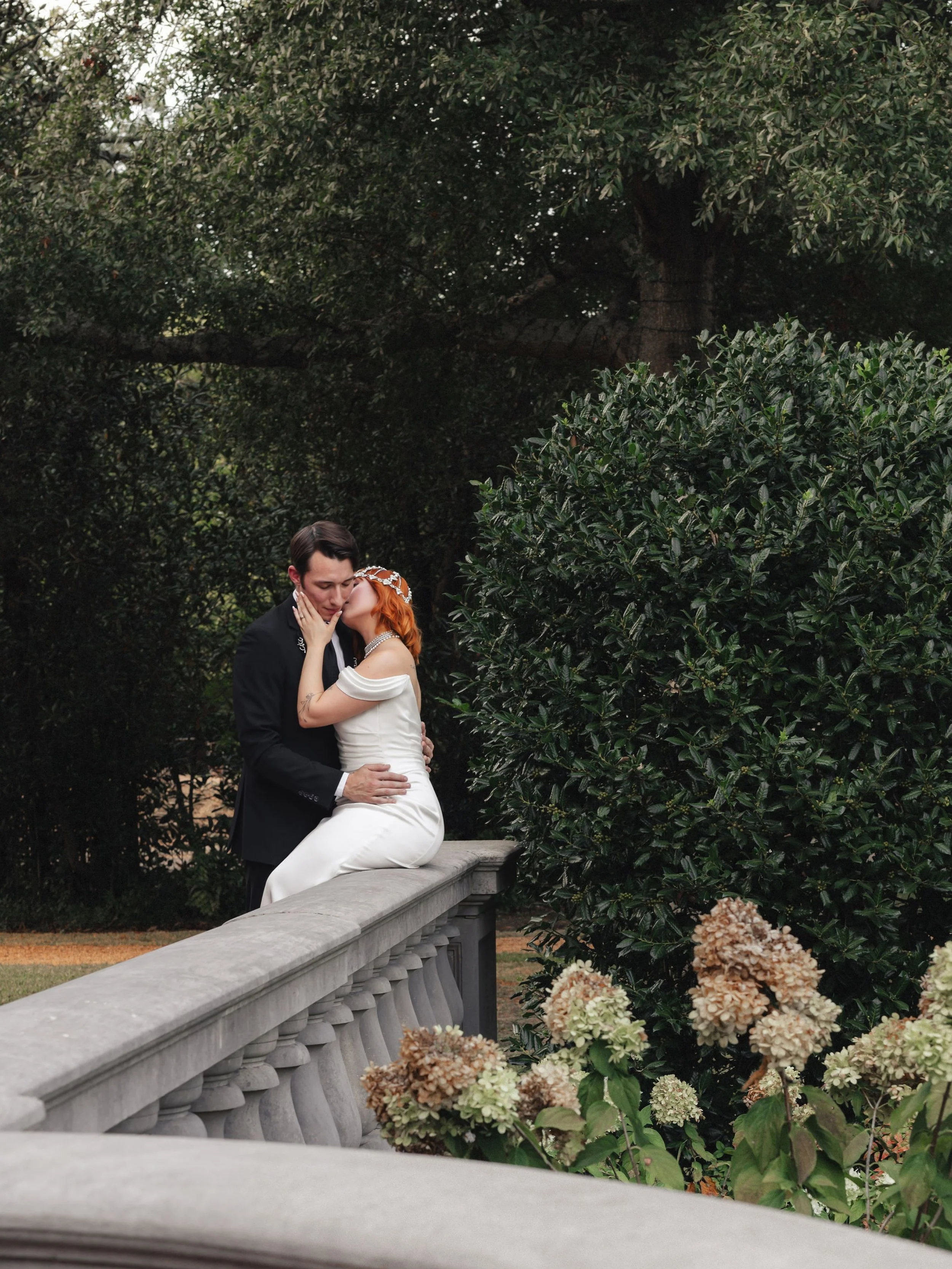 A couple dressed in wedding attire sharing a kiss outdoors near greenery and hydrangea flowers.