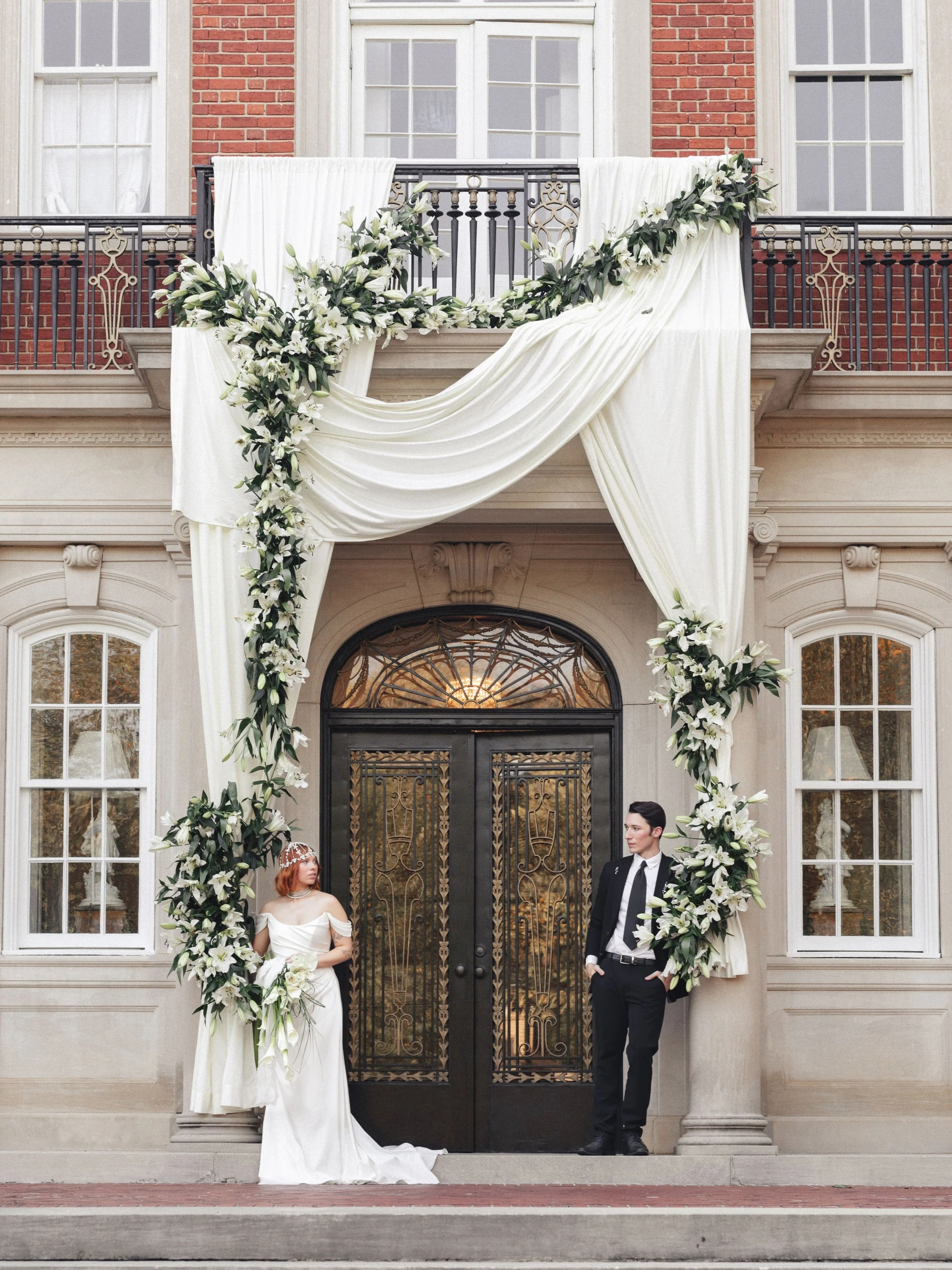 Bride in a white wedding gown holding a bouquet standing on the left, groom in a black suit leaning against a column on the right, elegant building entrance decorated with white drapes and flowers, large windows, and wrought-iron balcony above.