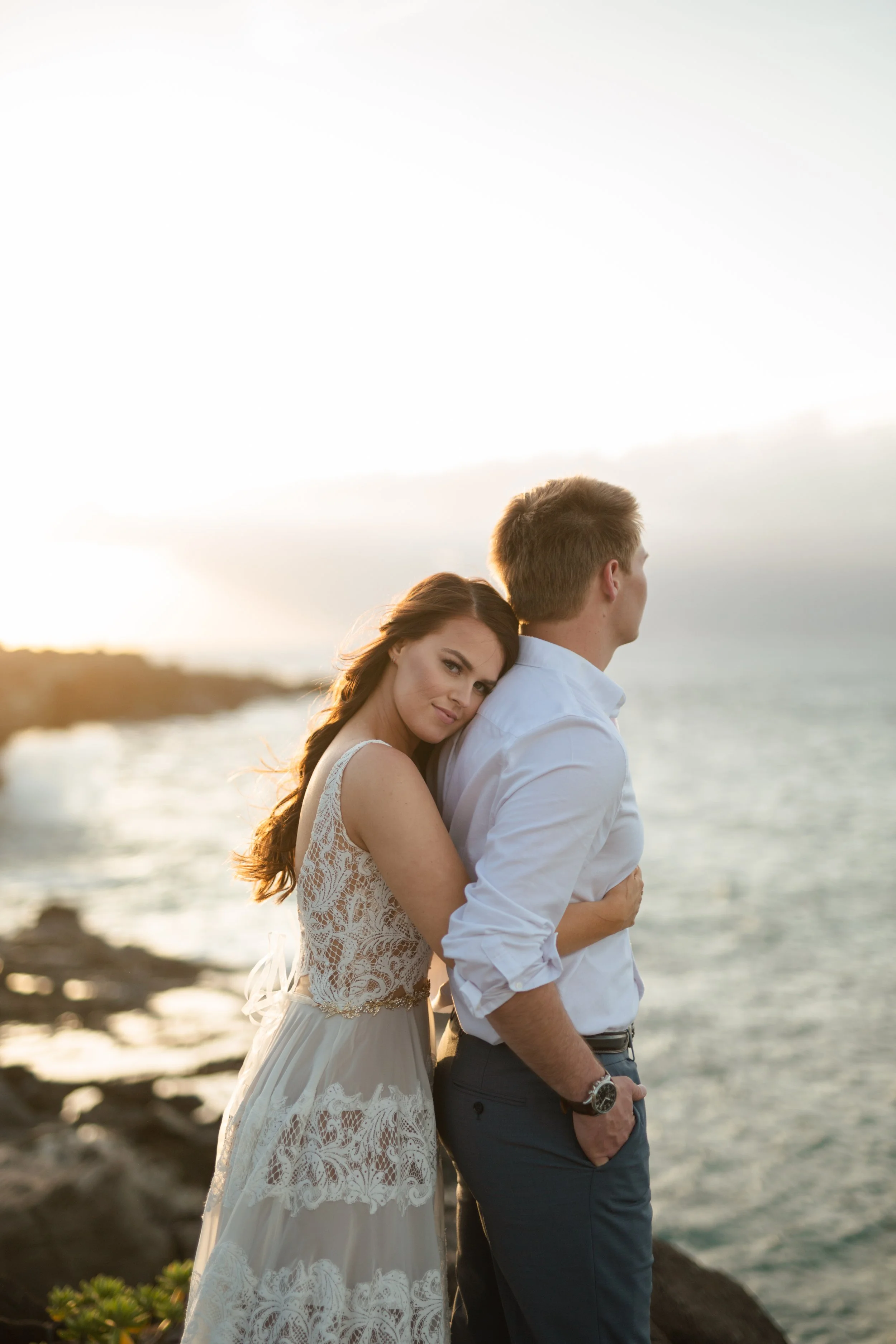 A woman and a man stand clos by the water during sunset; the woman leans her head on the man’s shoulder, and they both look into the distance. The woman wears a white lace dress, and the man wears a white shirt and dark pants.