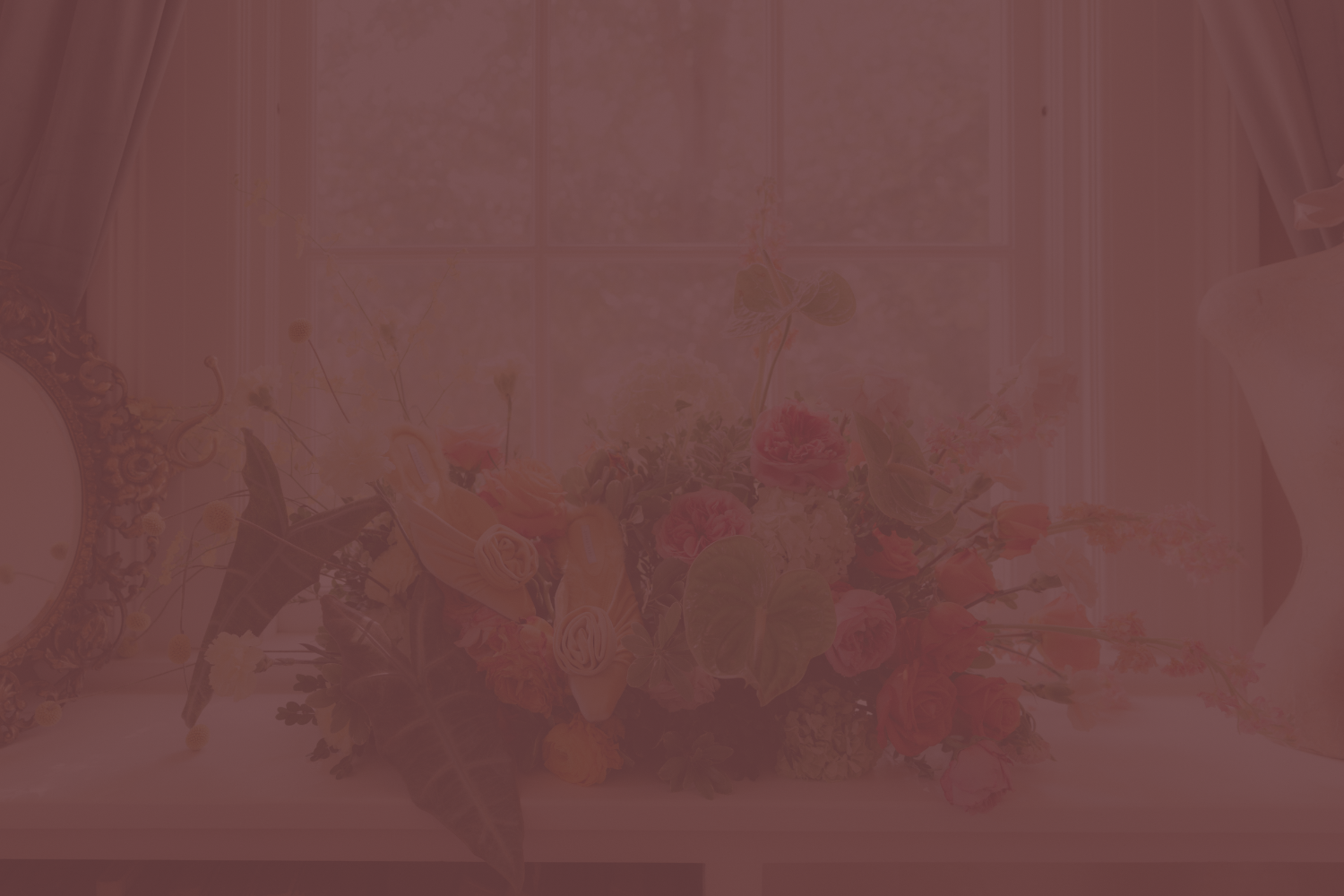 A table with a large colorful flower arrangement, a small clock, and a mirror in the background near a window with curtains.