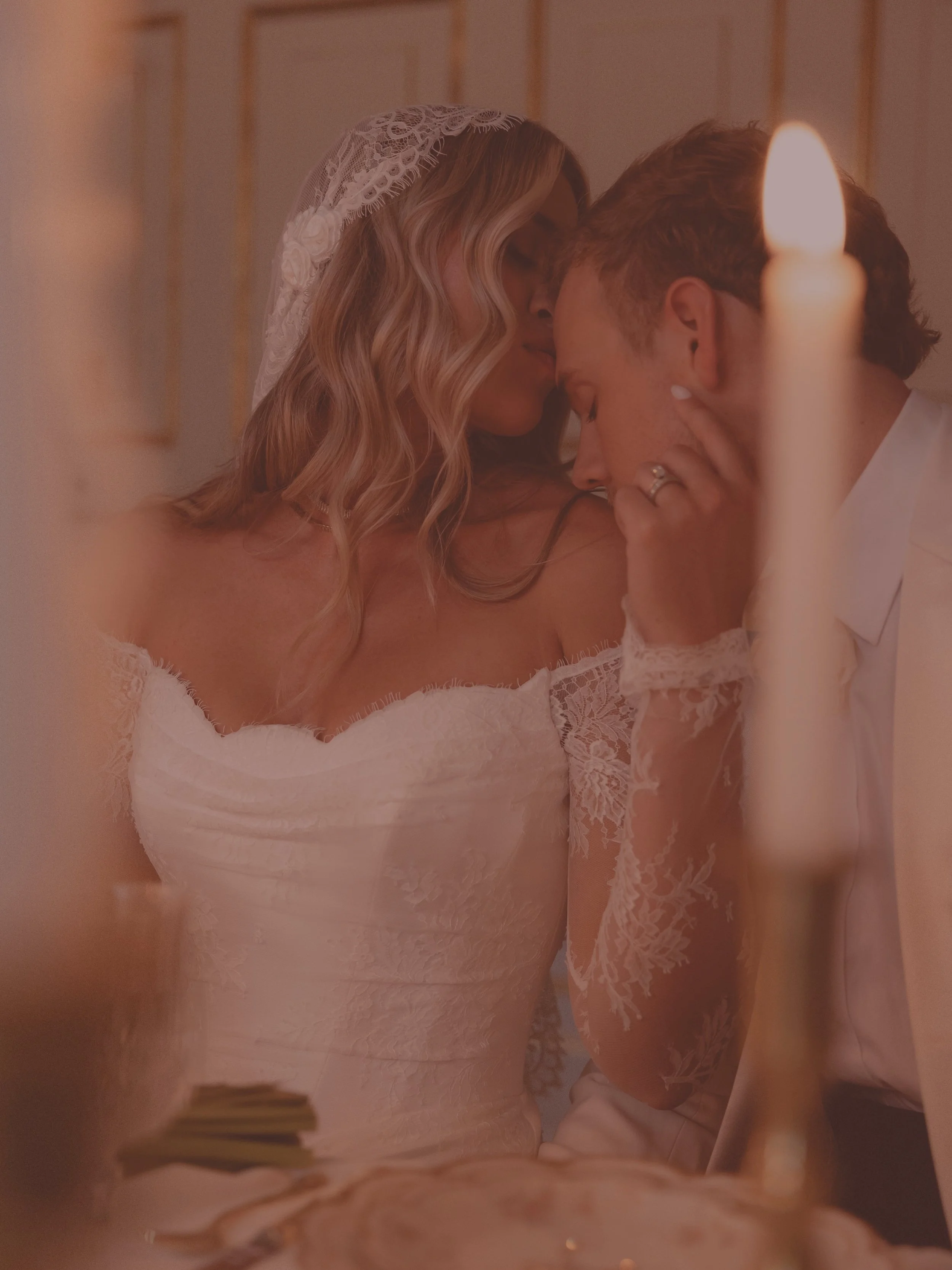 A bride and groom share an intimate moment during their wedding ceremony, with the bride wearing a lace wedding dress and veil, and the groom in a light-colored suit, with a candle in the foreground.